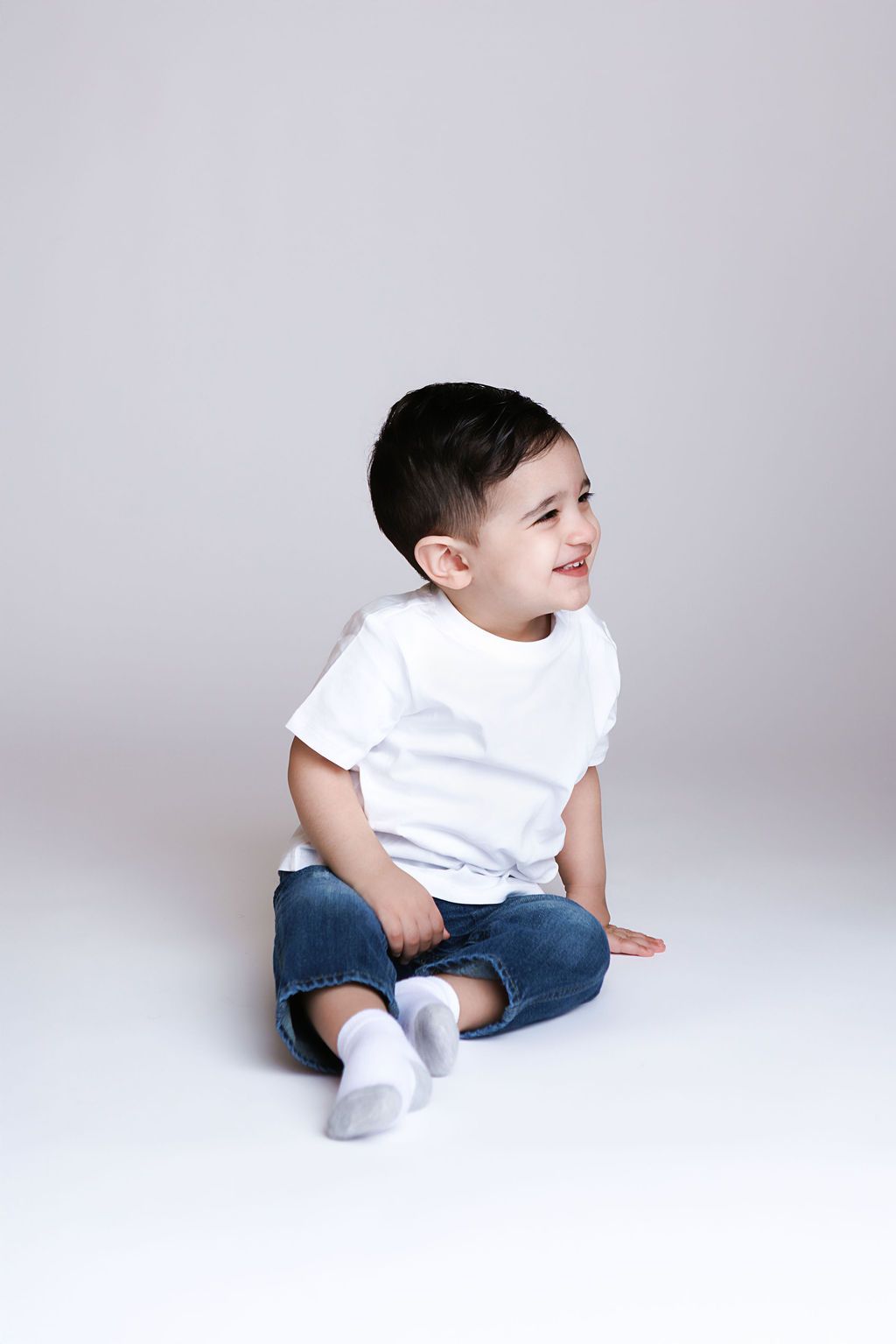 Smiling toddler boy sitting on white floor wearing white t-shirt, jeans, and white socks.