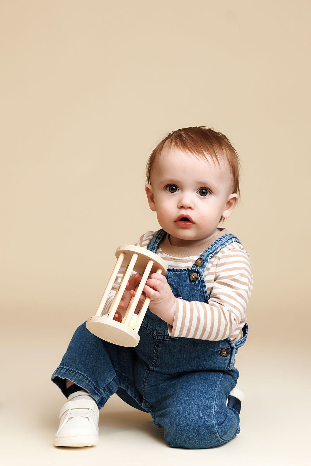 Baby in denim overalls and striped shirt sits on floor holding a wooden toy rattle.
