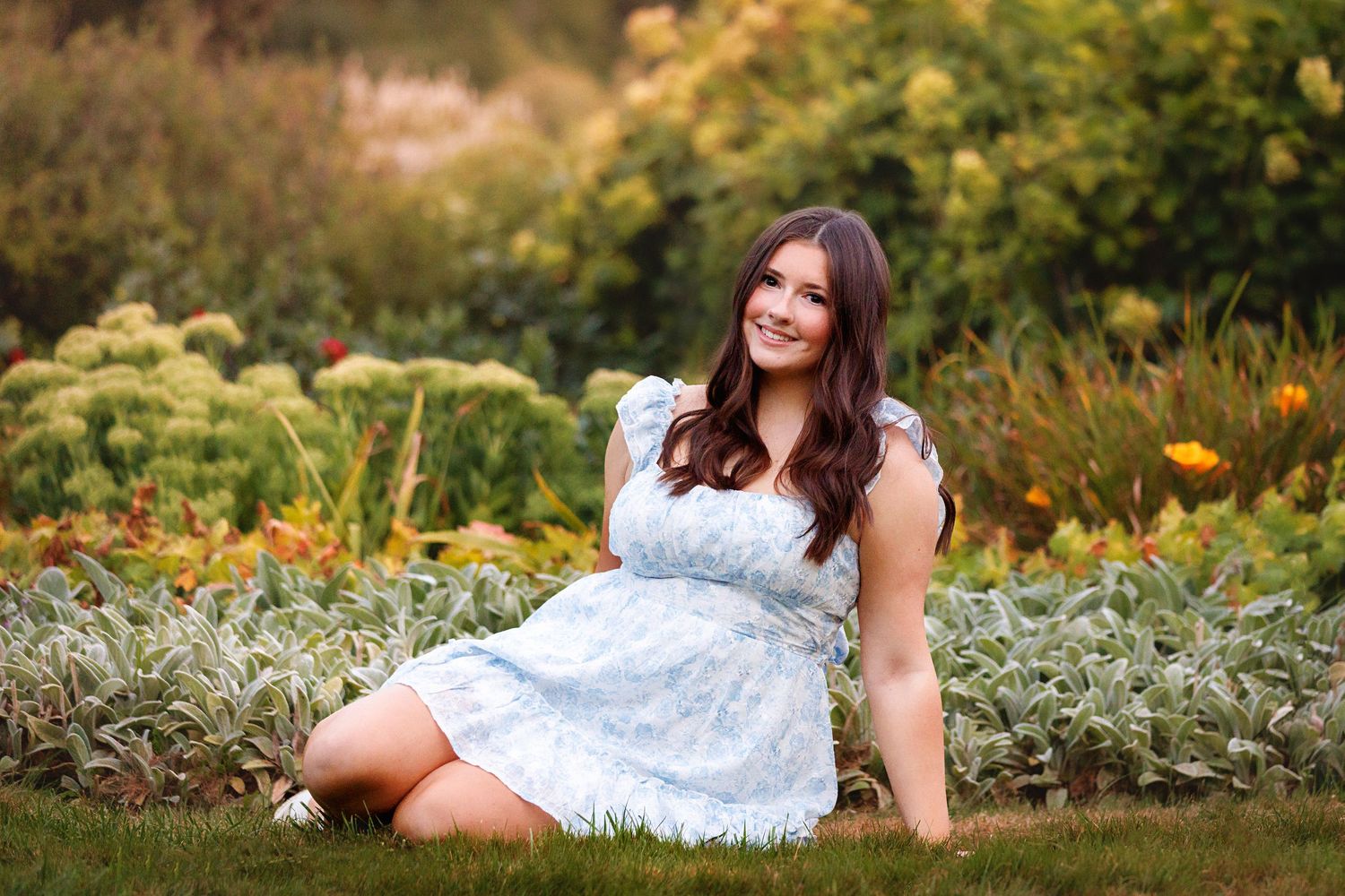 Young woman in light blue floral dress sitting on grass in a colorful garden, smiling outdoors.