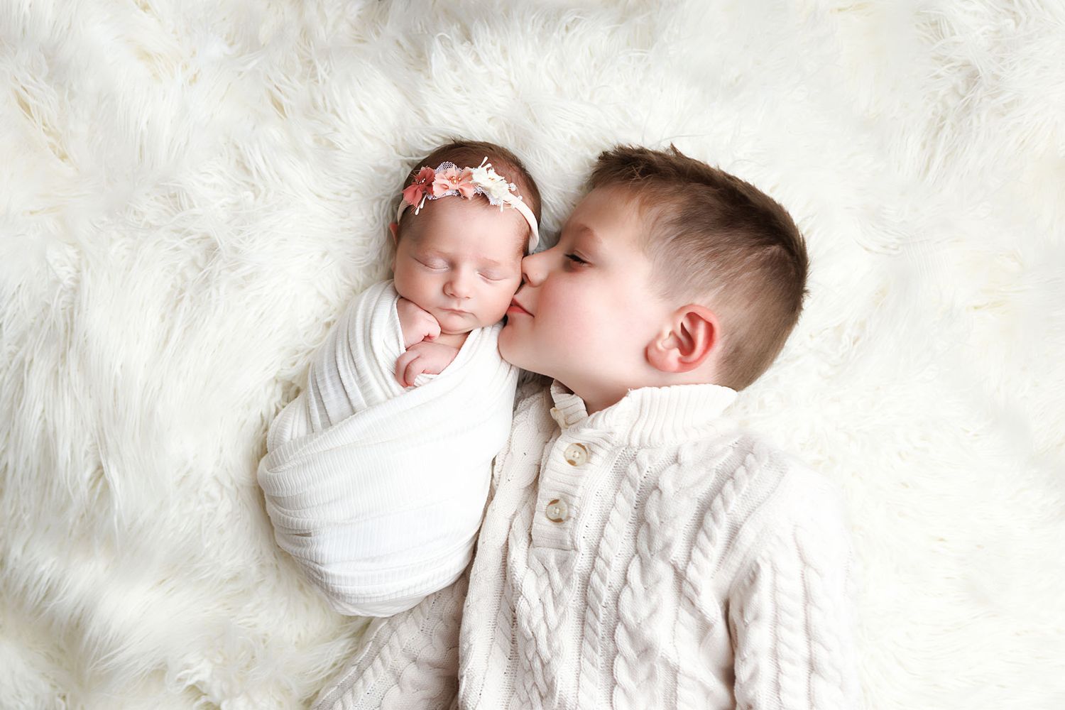Older boy in white knit sweater gently kissing sleeping newborn baby sister wrapped in white on fluffy fur blanket.