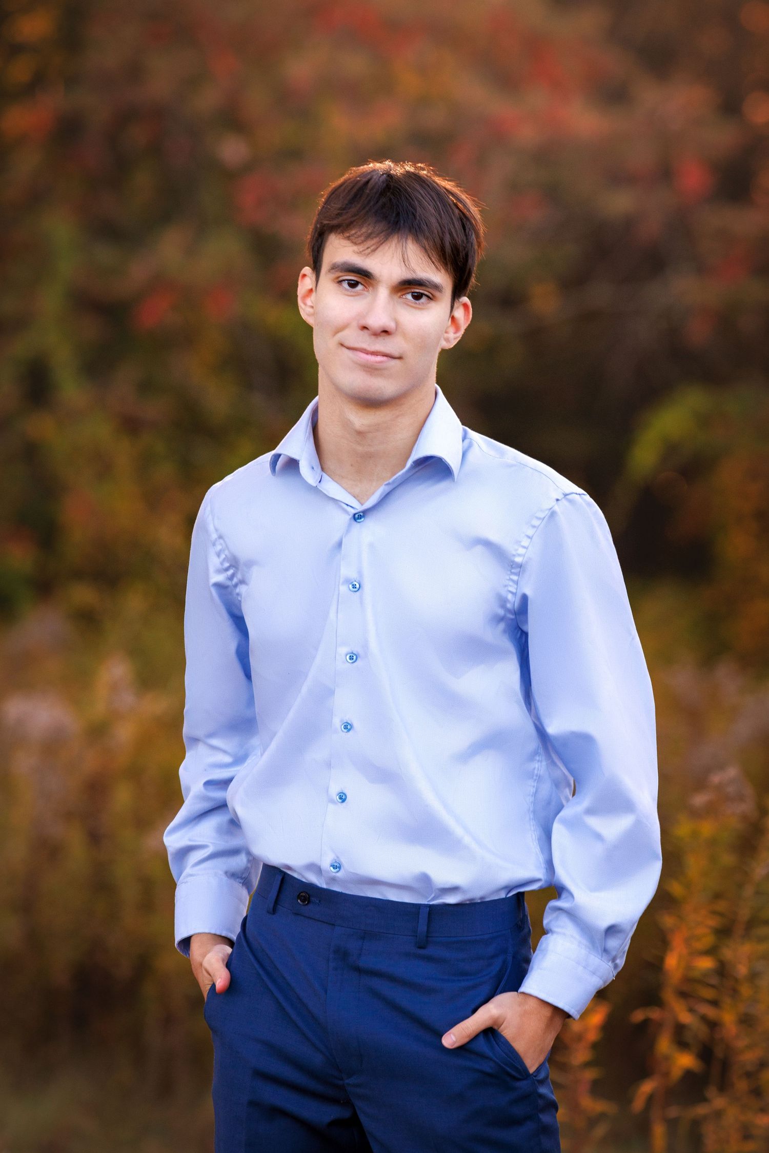 High School Senior in a light blue dress shirt and navy pants poses outdoors against an autumn background with red foliage.