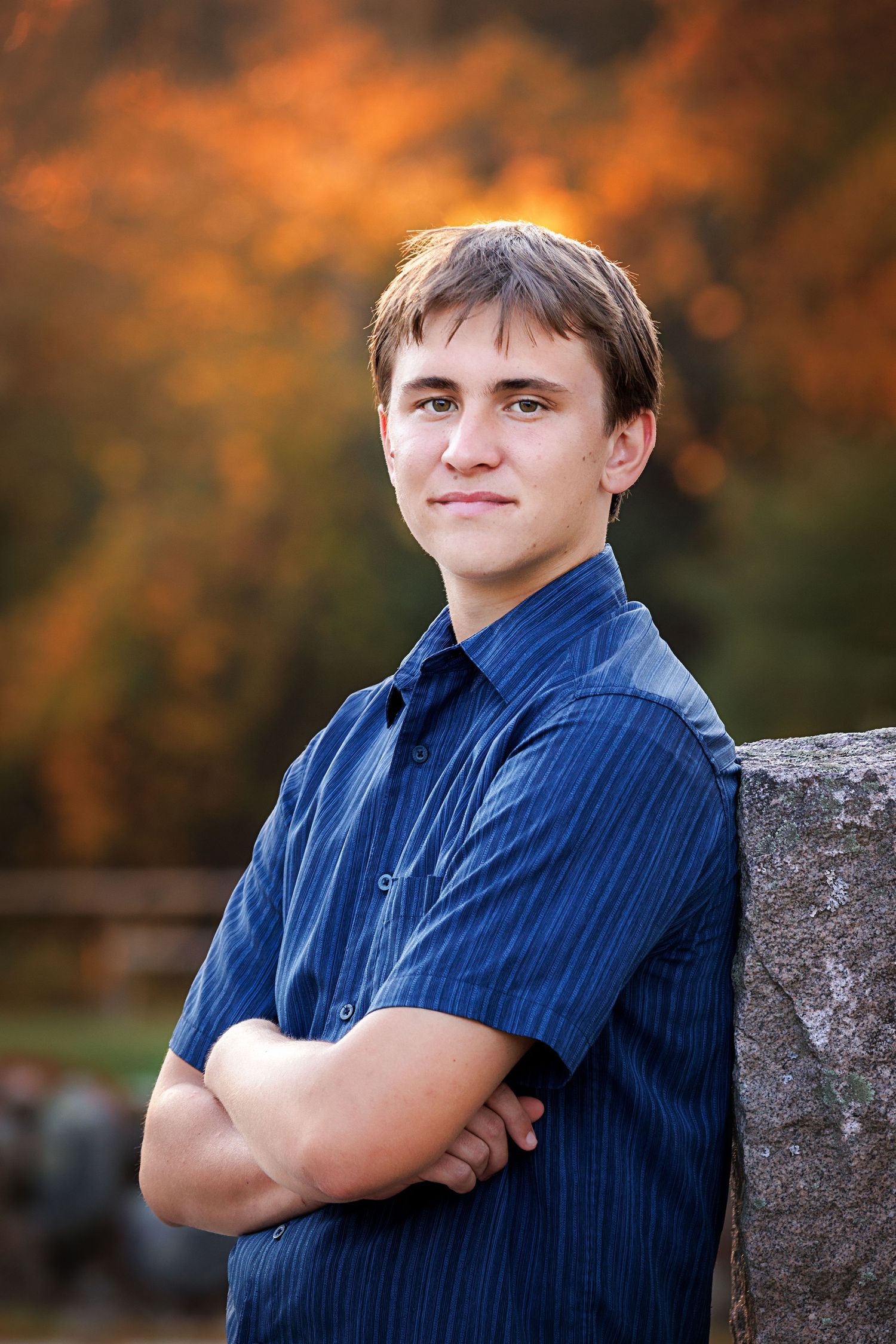 Portrait of a High School Senior in a blue striped shirt leaning against a stone wall with autumn foliage in the background.