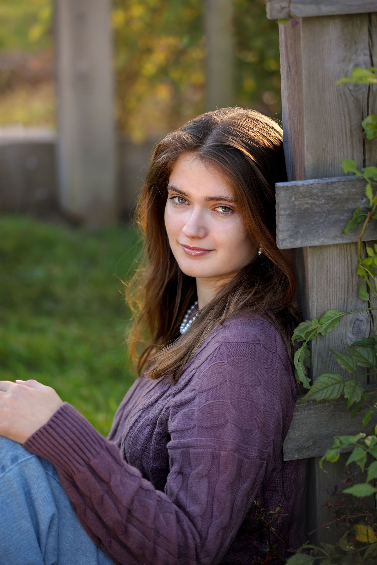 A High School Senior in a purple sweater sits against a wooden post outdoors during golden hour lighting.