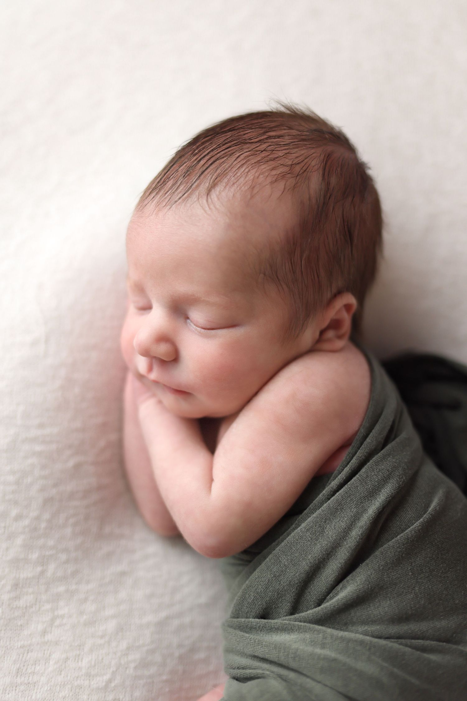 A sleeping newborn baby wrapped in a sage green swaddle blanket against a white textured background.