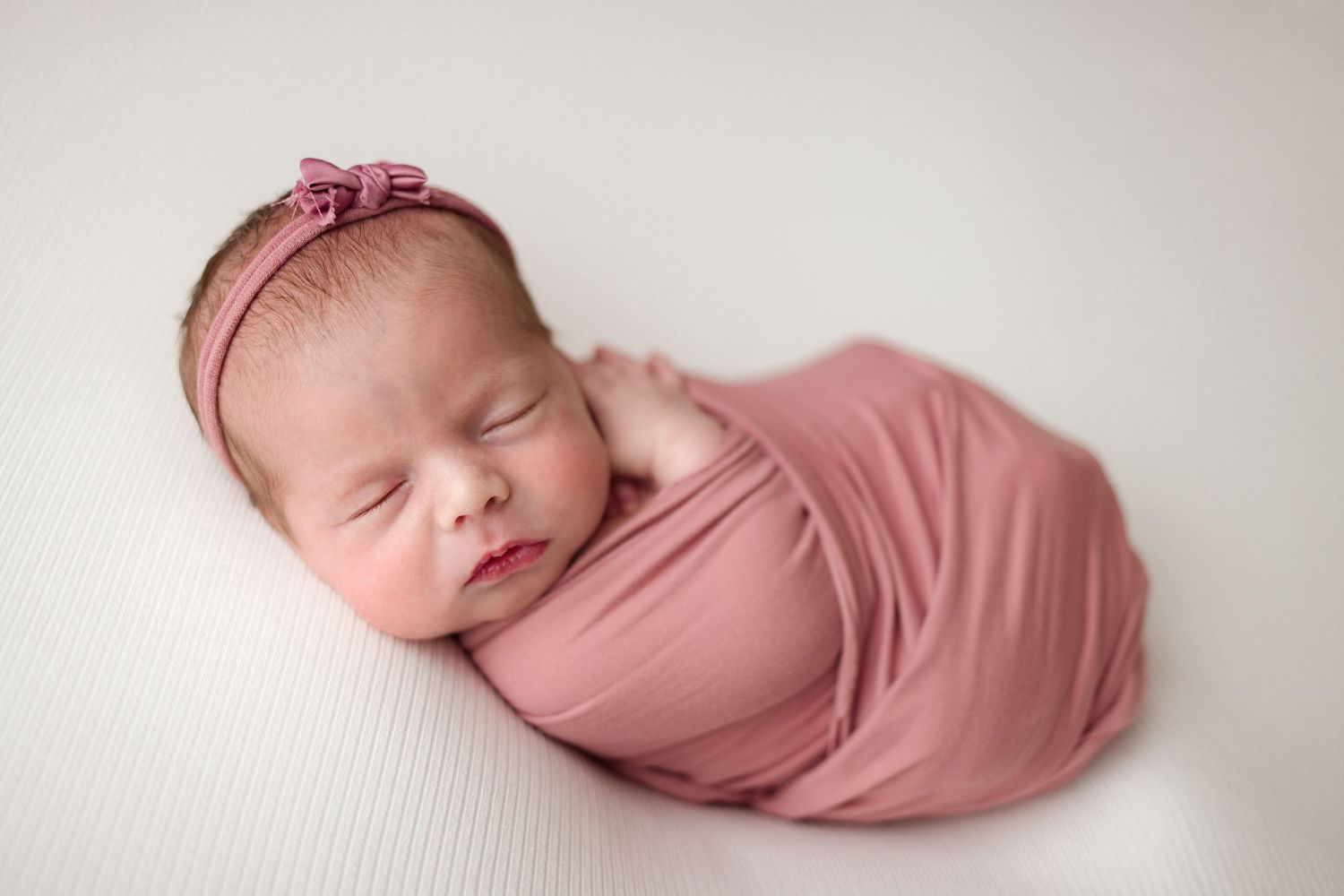 Peaceful sleeping newborn baby wrapped in soft dusty rose fabric against white background.