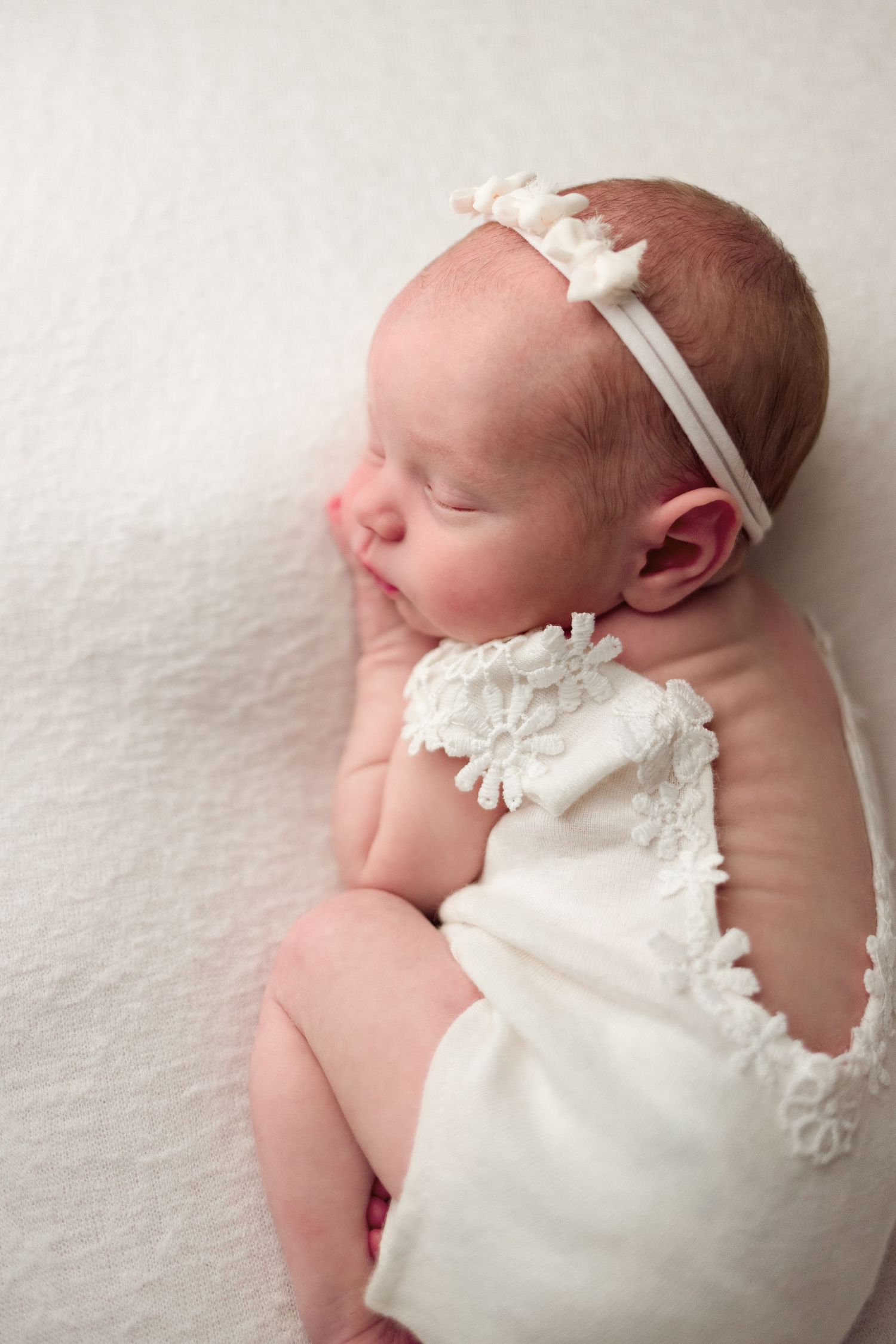 Sleeping newborn baby wearing a delicate white dress and headband lies peacefully on soft white bedding.