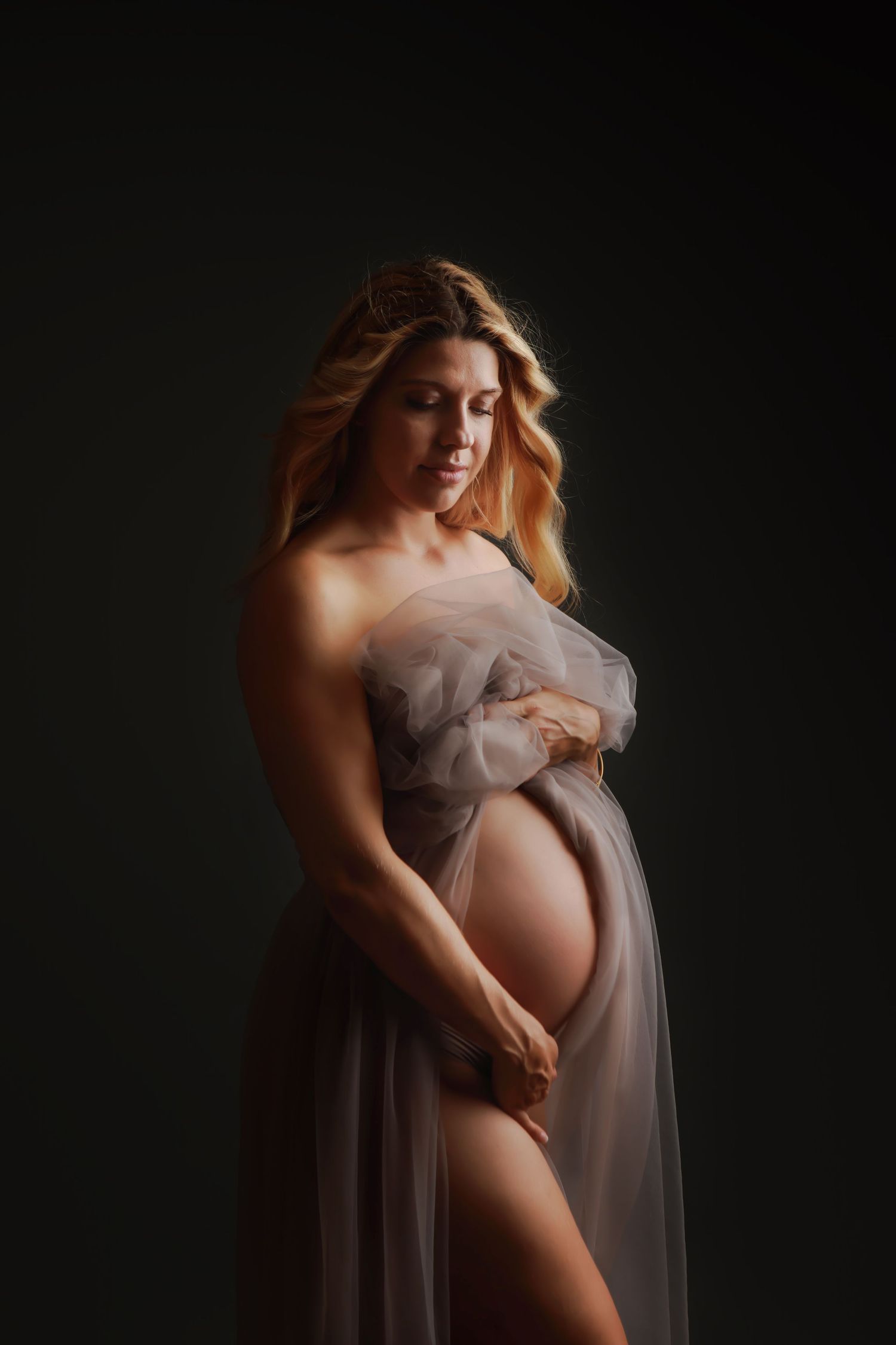 Artistic maternity portrait with flowing sheer fabric against dark background in dramatic lighting.
