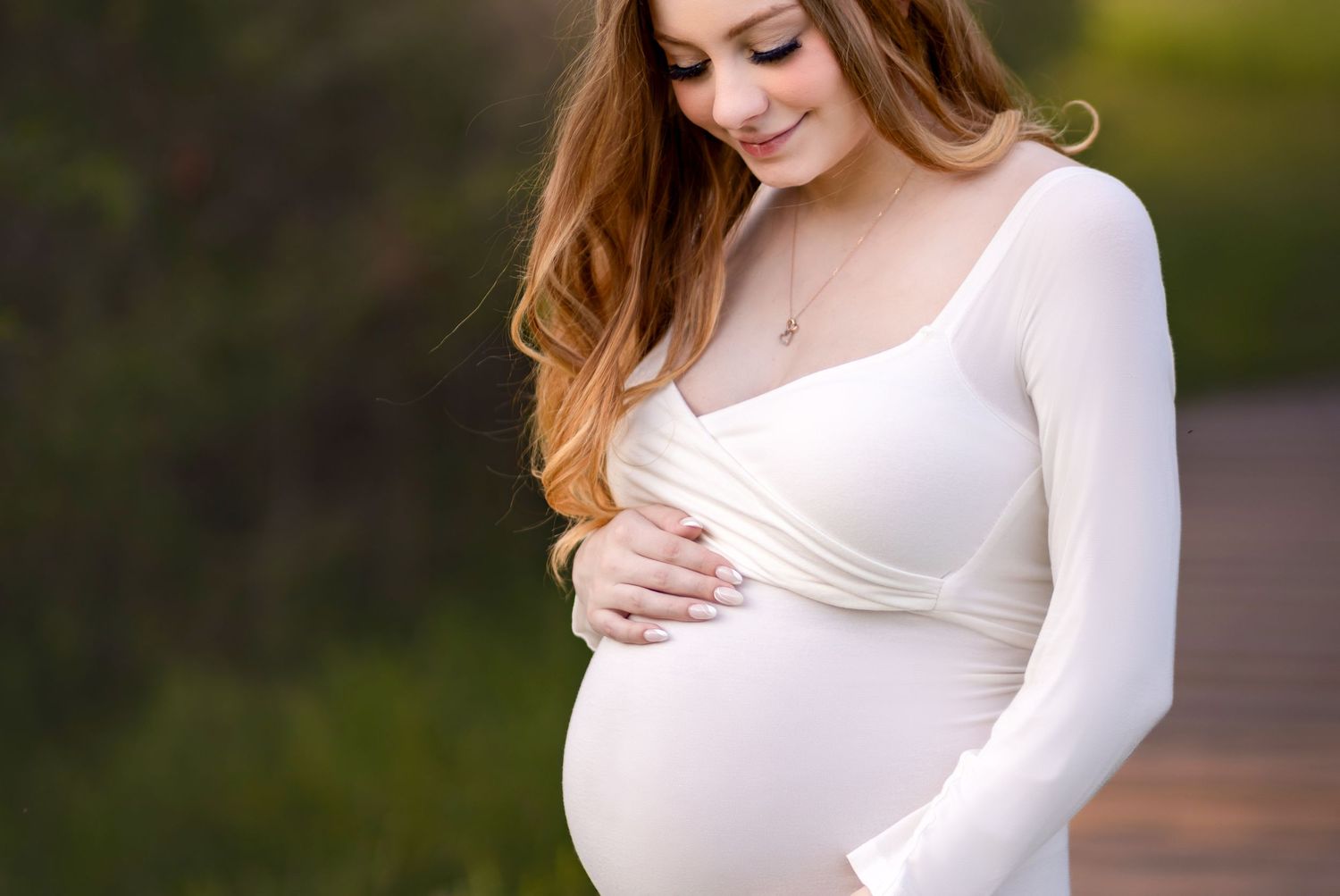 Expecting mother in white dress caresses baby bump while standing outdoors in soft natural lighting.