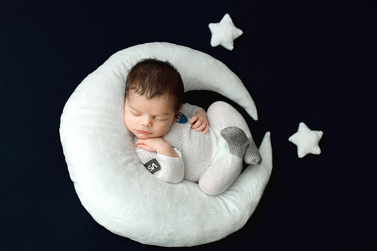 A sleeping newborn baby rests peacefully on a white crescent moon prop with small stars in the background.