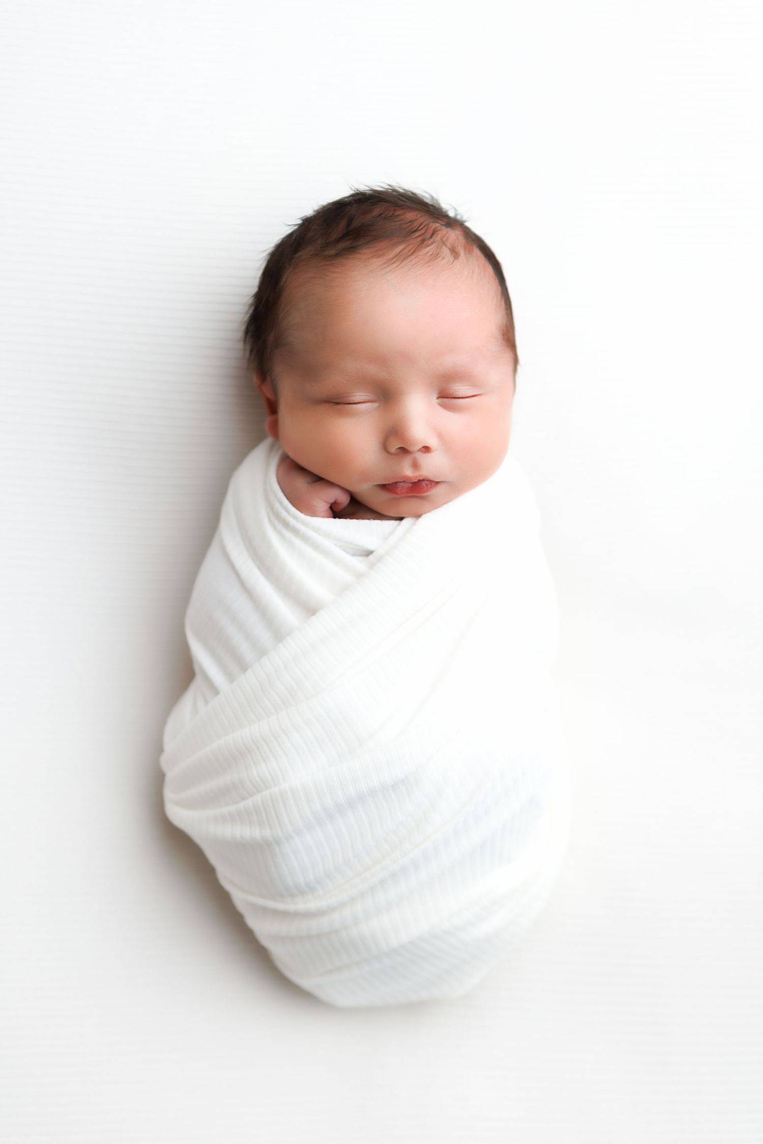 Sleeping newborn baby swaddled in a white wrap blanket, lying on a white background.