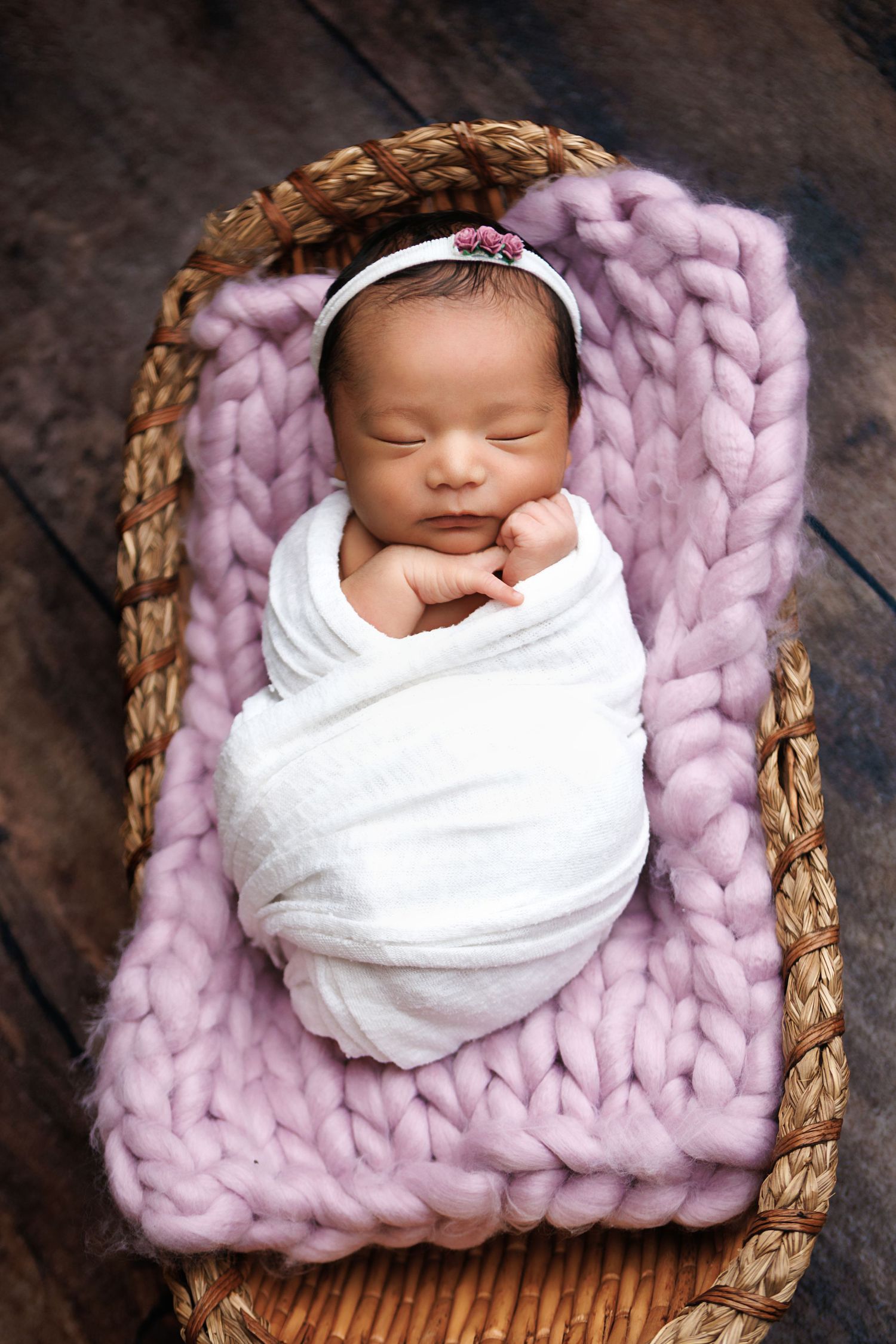 Sleeping newborn baby swaddled in white wrap with pink headband resting in wicker basket on chunky knit blanket.