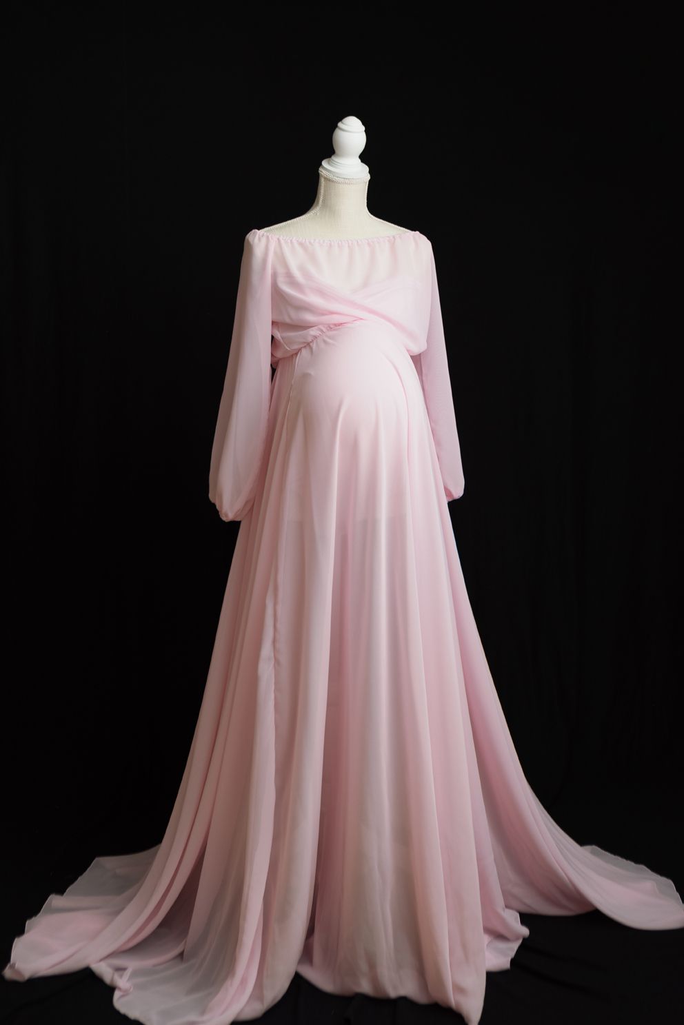 Elegant pink chiffon maternity gown with long sleeves and flowing train displayed on white mannequin against black backdrop.