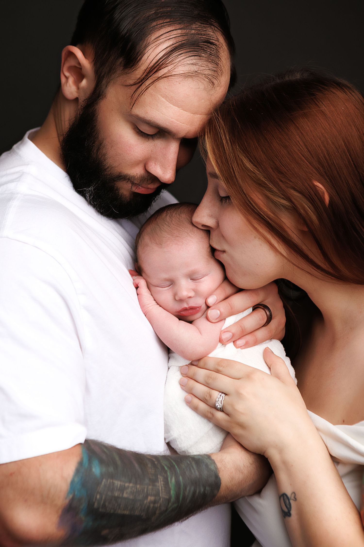 Loving parents gently holding and kissing their sleeping newborn baby, father with beard and tattoos, mother with red hair.