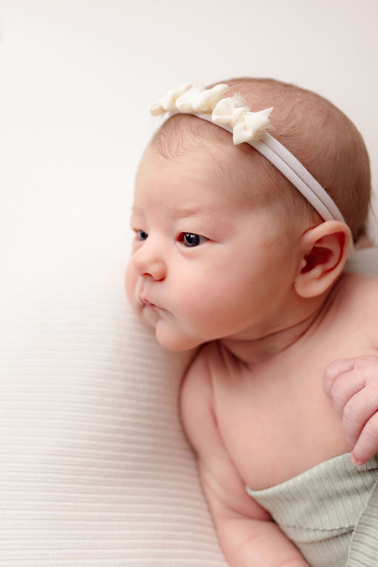 Newborn baby wearing mint green outfit and white headband rests peacefully on white textured blanket.