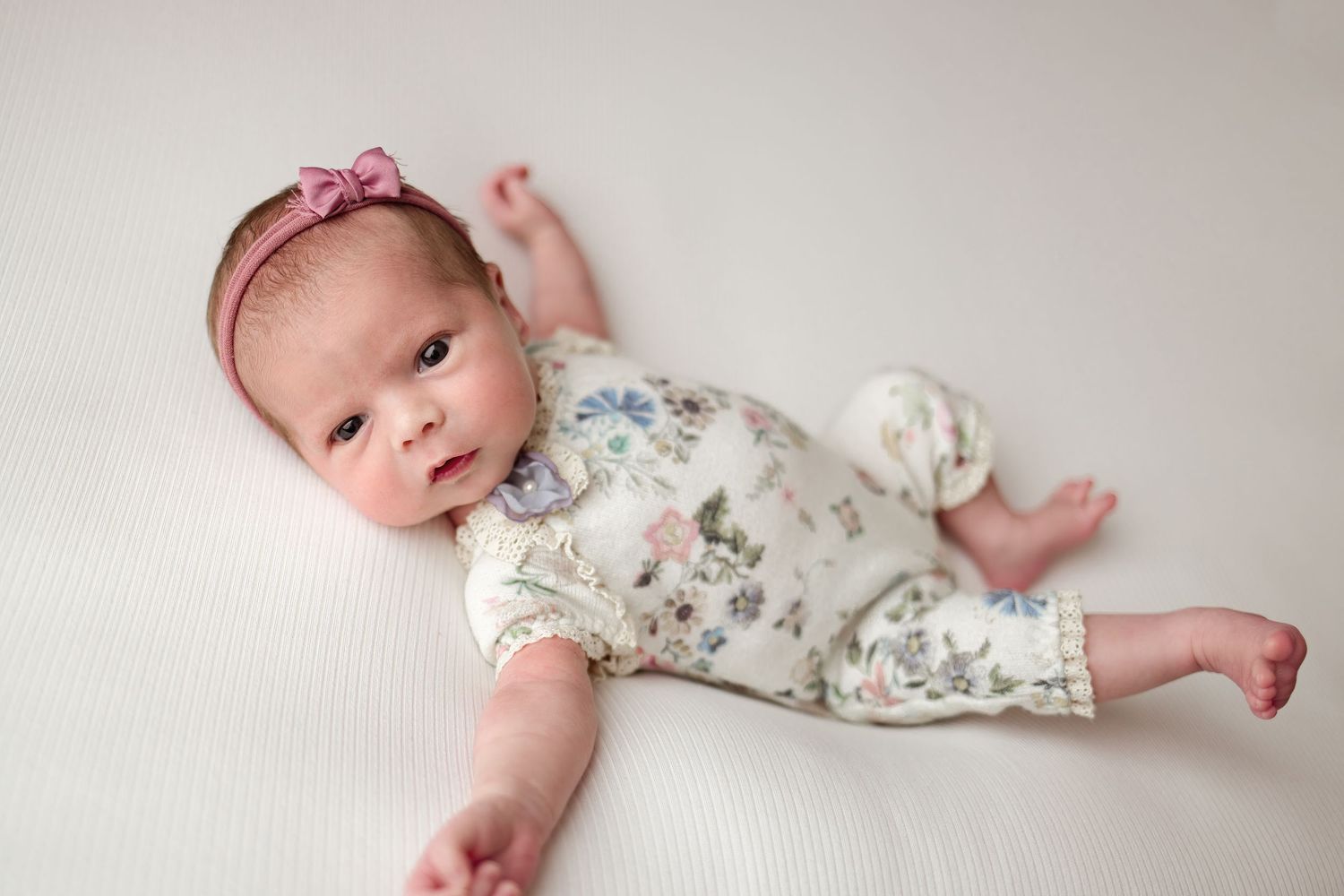 Baby wearing floral romper and pink bow headband lying on white textured backdrop during newborn photo session.