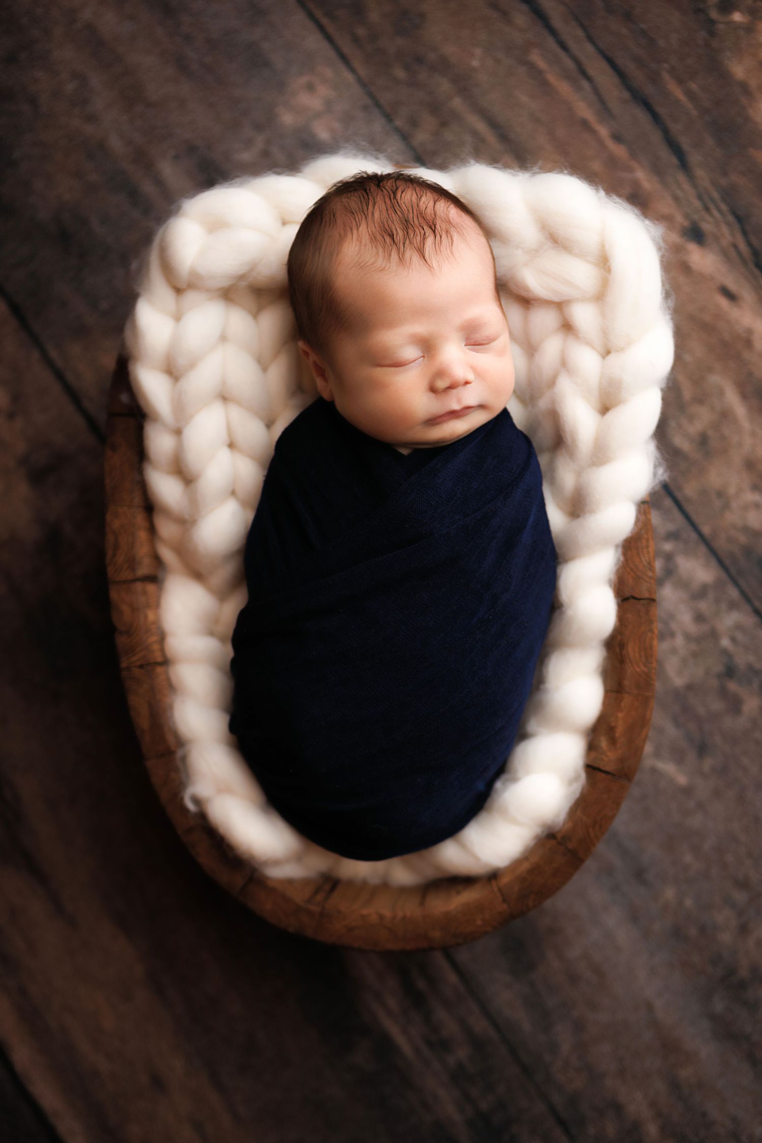 A sleeping newborn wrapped in navy blue swaddle rests peacefully in a rustic wooden bowl filled with fluffy white wool.
