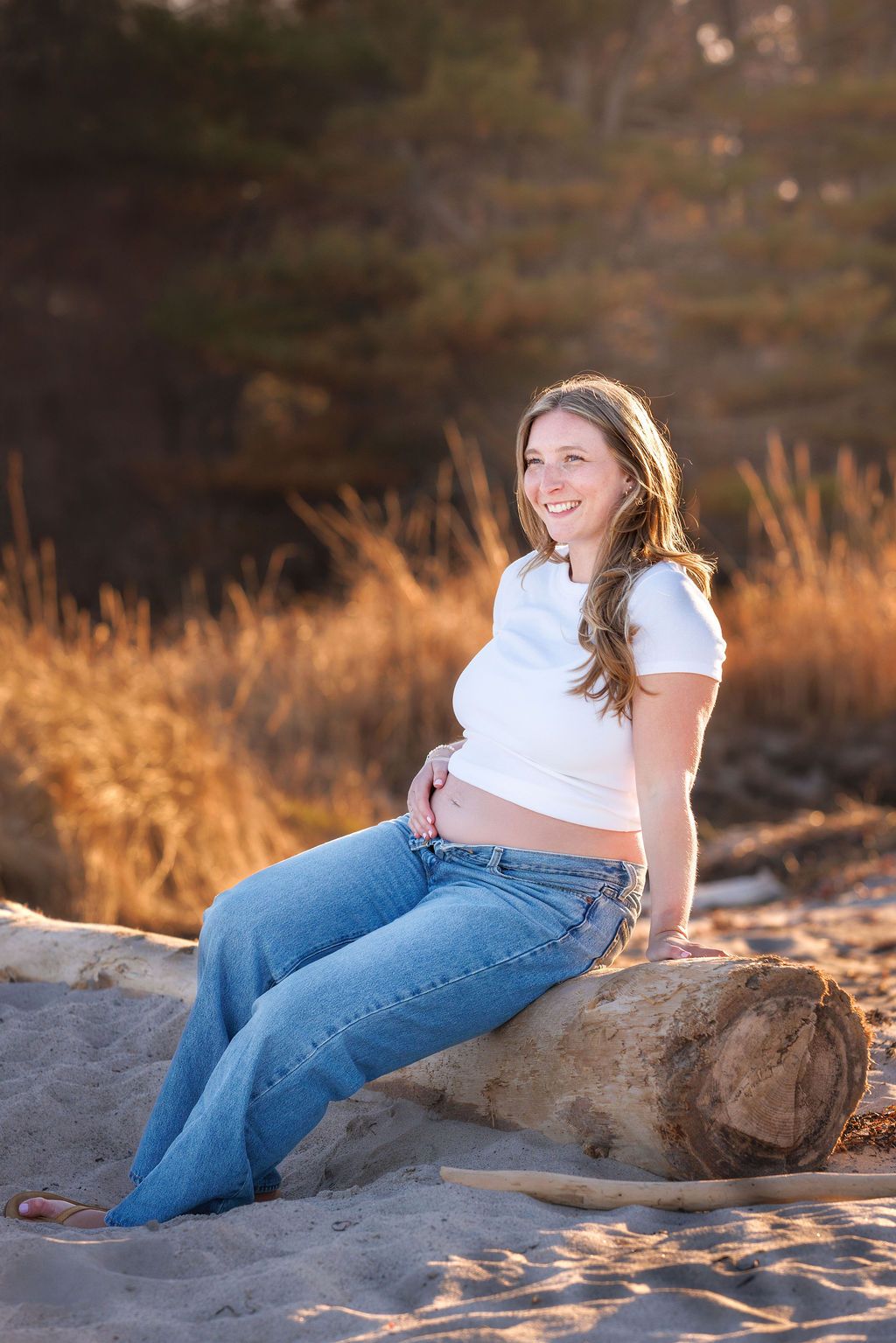 Smiling pregnant woman in white crop top and jeans sitting on driftwood log at sandy beach during golden hour.