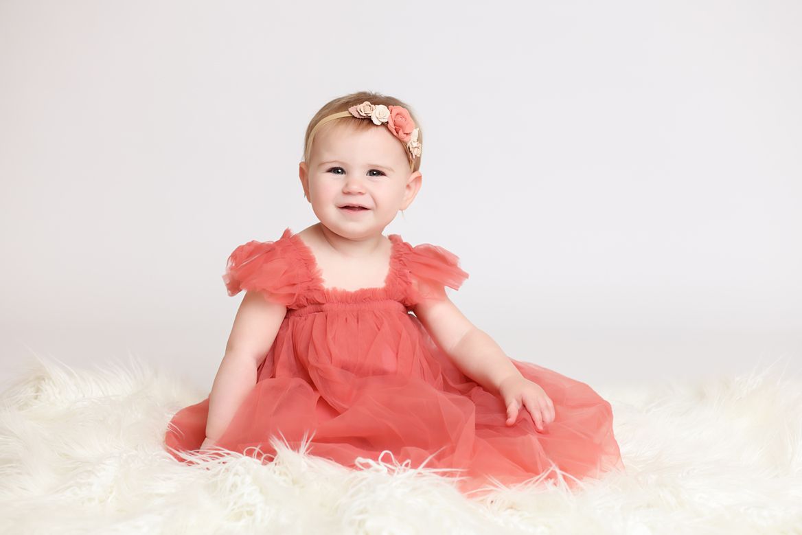 Baby in coral pink dress sitting and crawling on white backdrop with balloons.