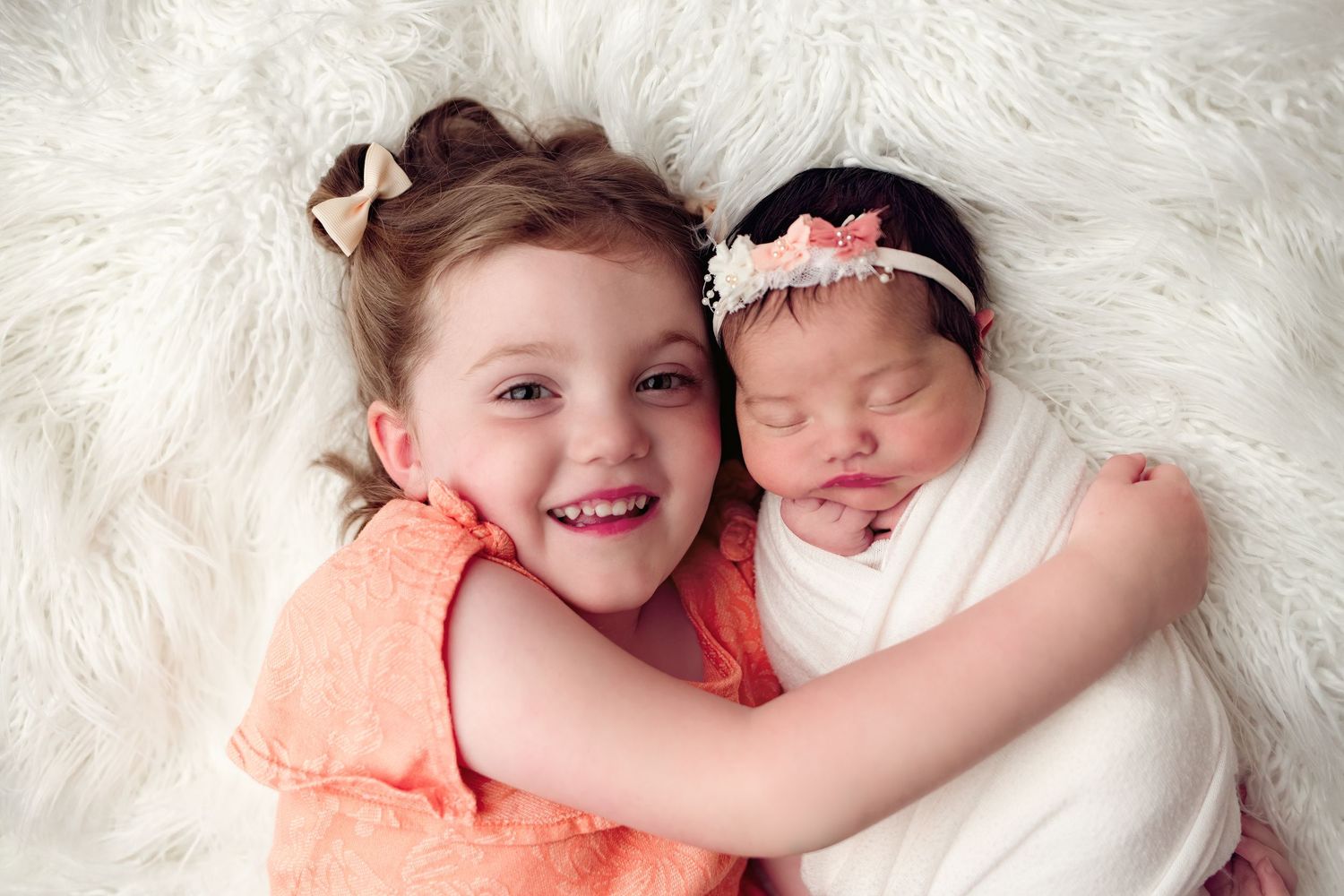 Two children lying on white fur, one in a coral dress and one swaddled in white with a headband.