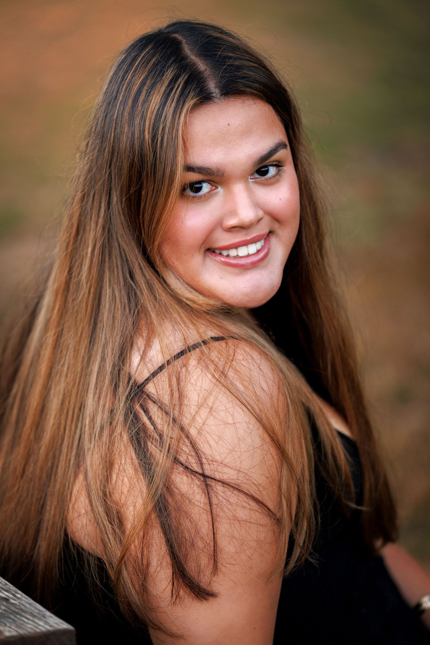 Portrait of High School Senior with long brown hair and a bright smile wearing black, photographed against a blurred natural background.