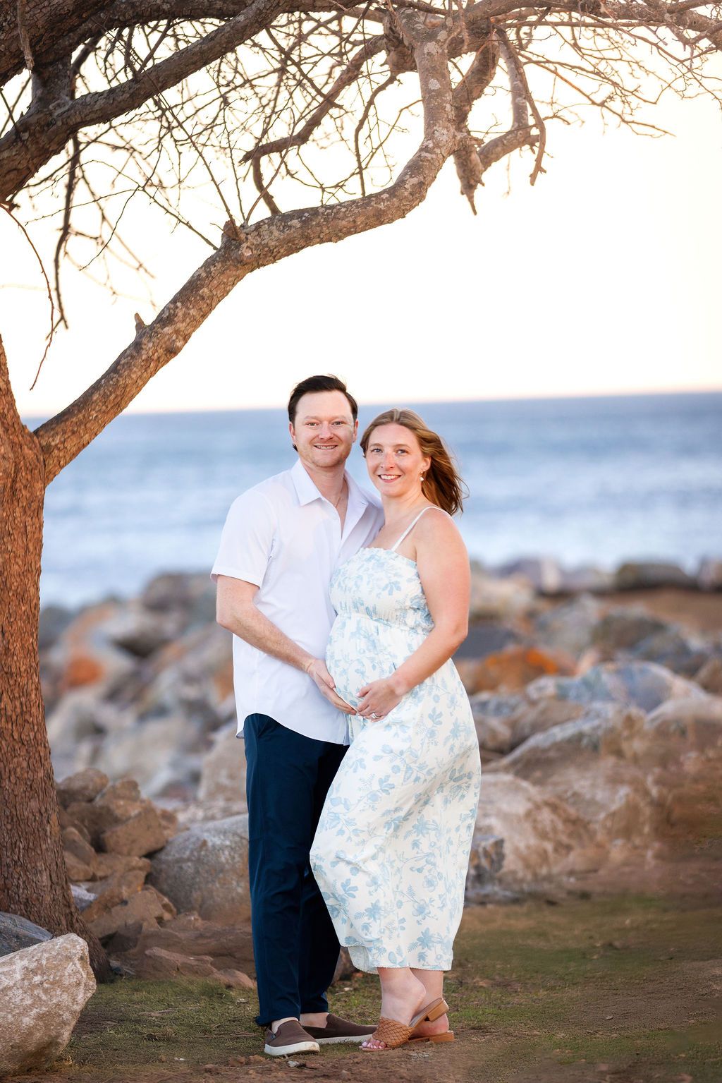 Pregnant couple embracing under a bare tree by rocky oceanside at sunset, woman in floral dress.