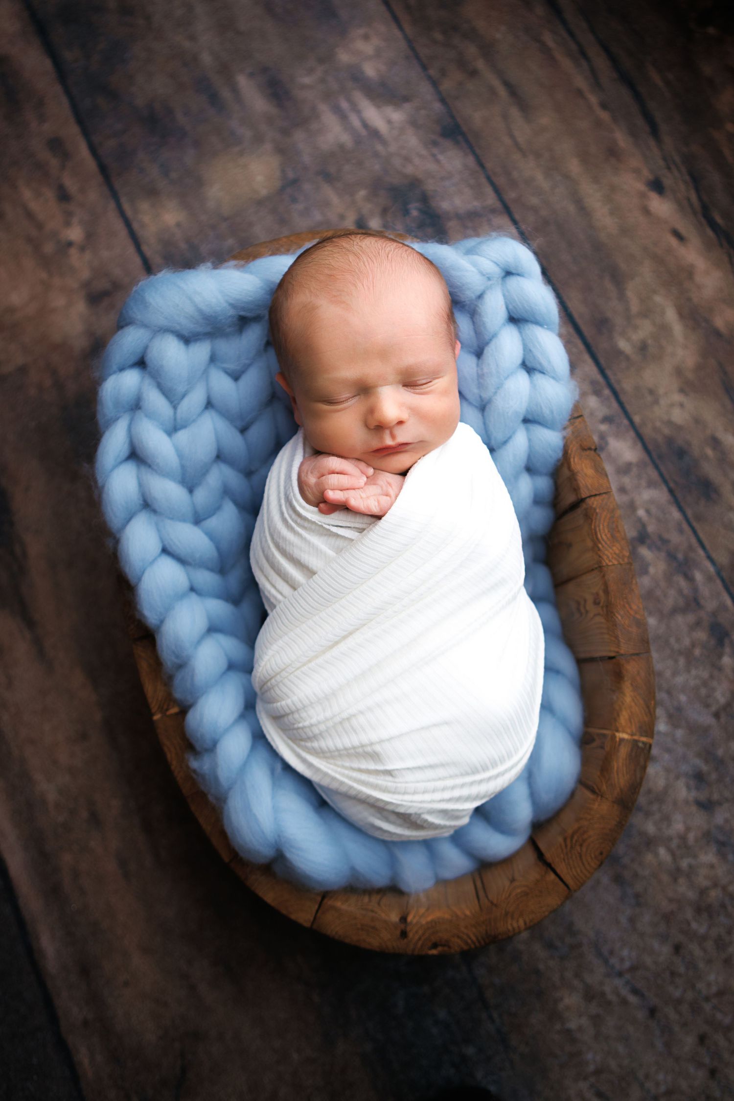 Newborn baby swaddled in white wrap sleeping peacefully in wooden bowl with blue chunky knit blanket.