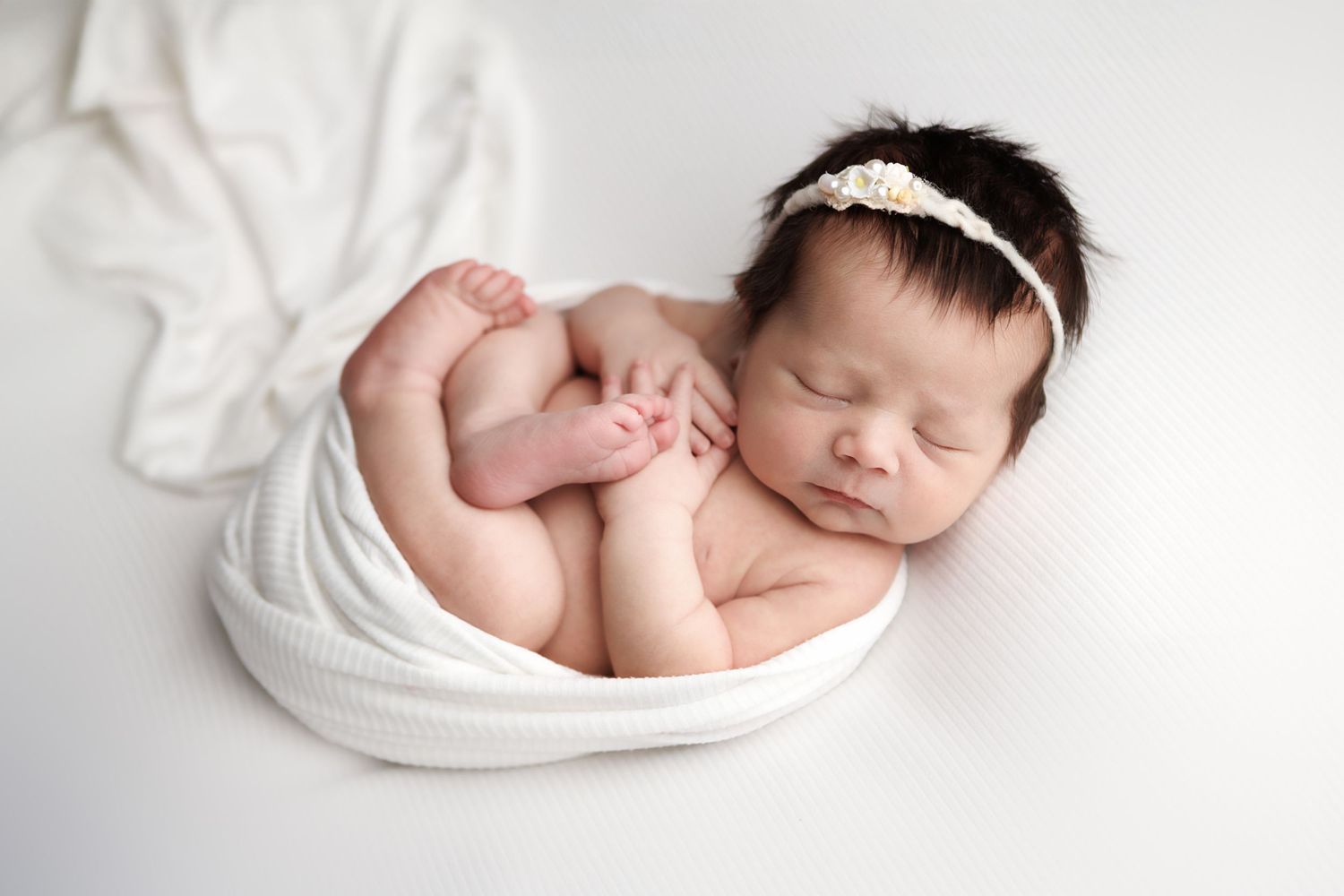 Sleeping newborn baby wrapped in white fabric wearing a headband while curled up in a white bowl.