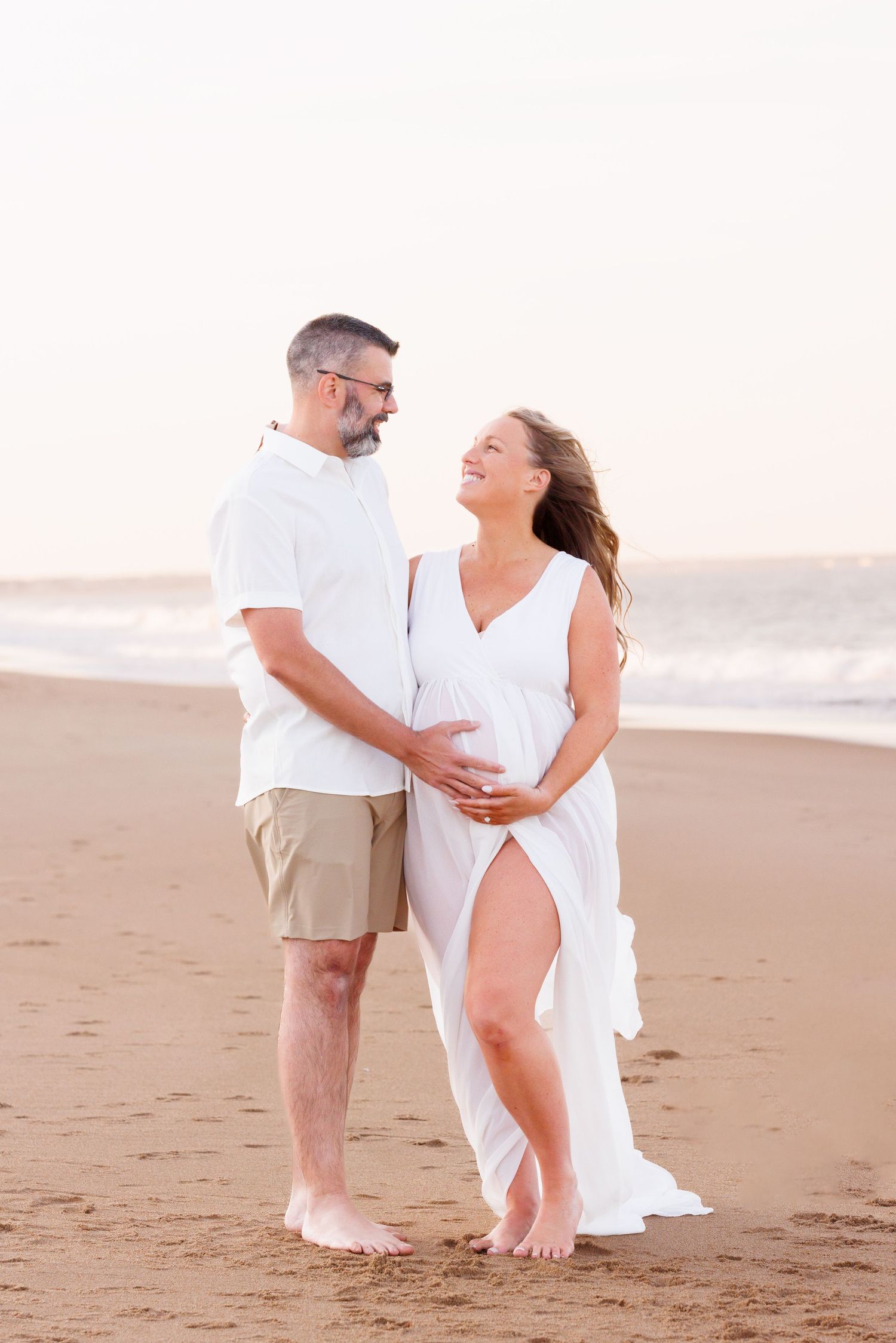 Couple sharing a joyful maternity moment on the beach, woman in white dress cradling baby bump, man in white shirt and khaki shorts.