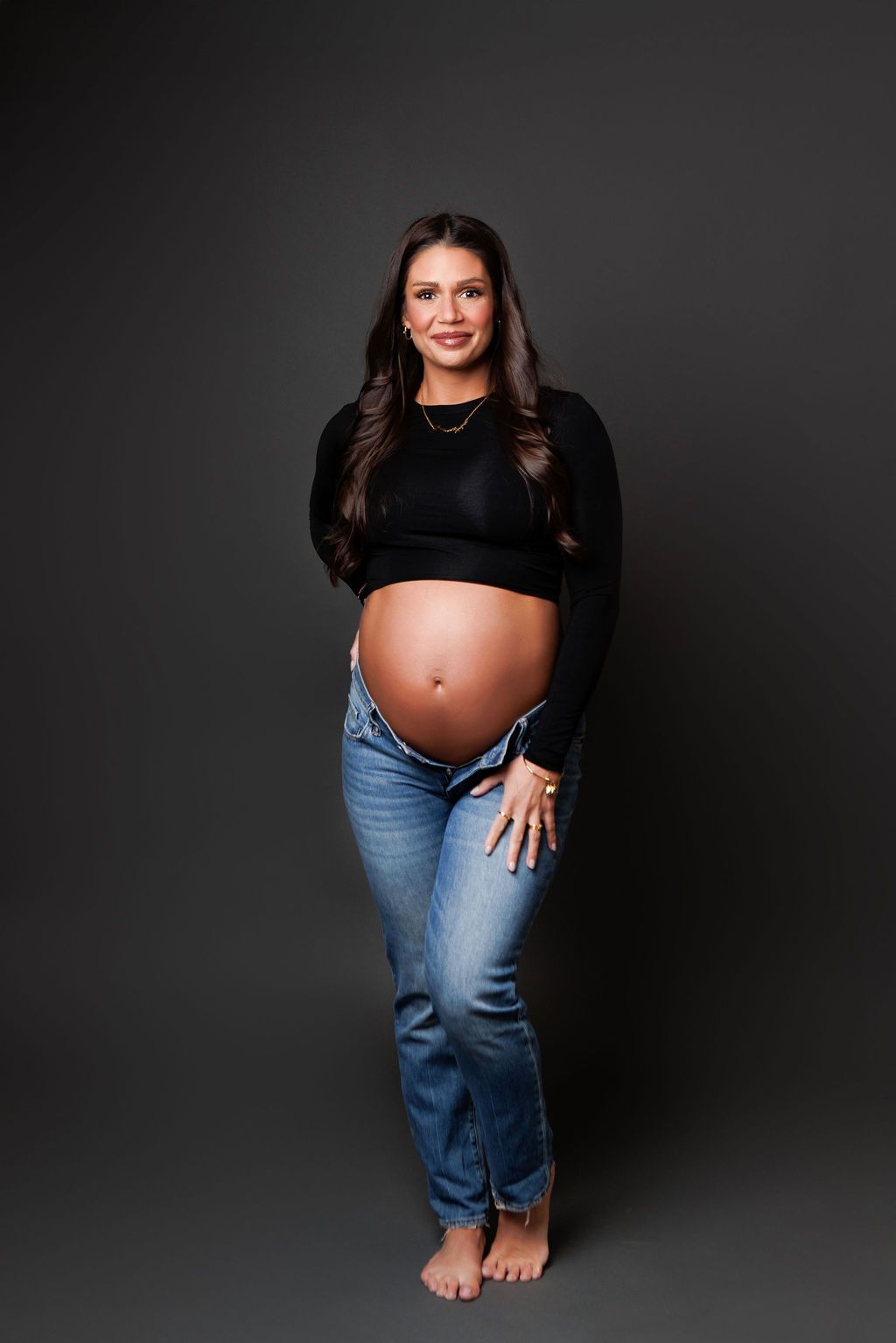 Smiling pregnant woman in black crop top and jeans posing barefoot against dark gray studio background.
