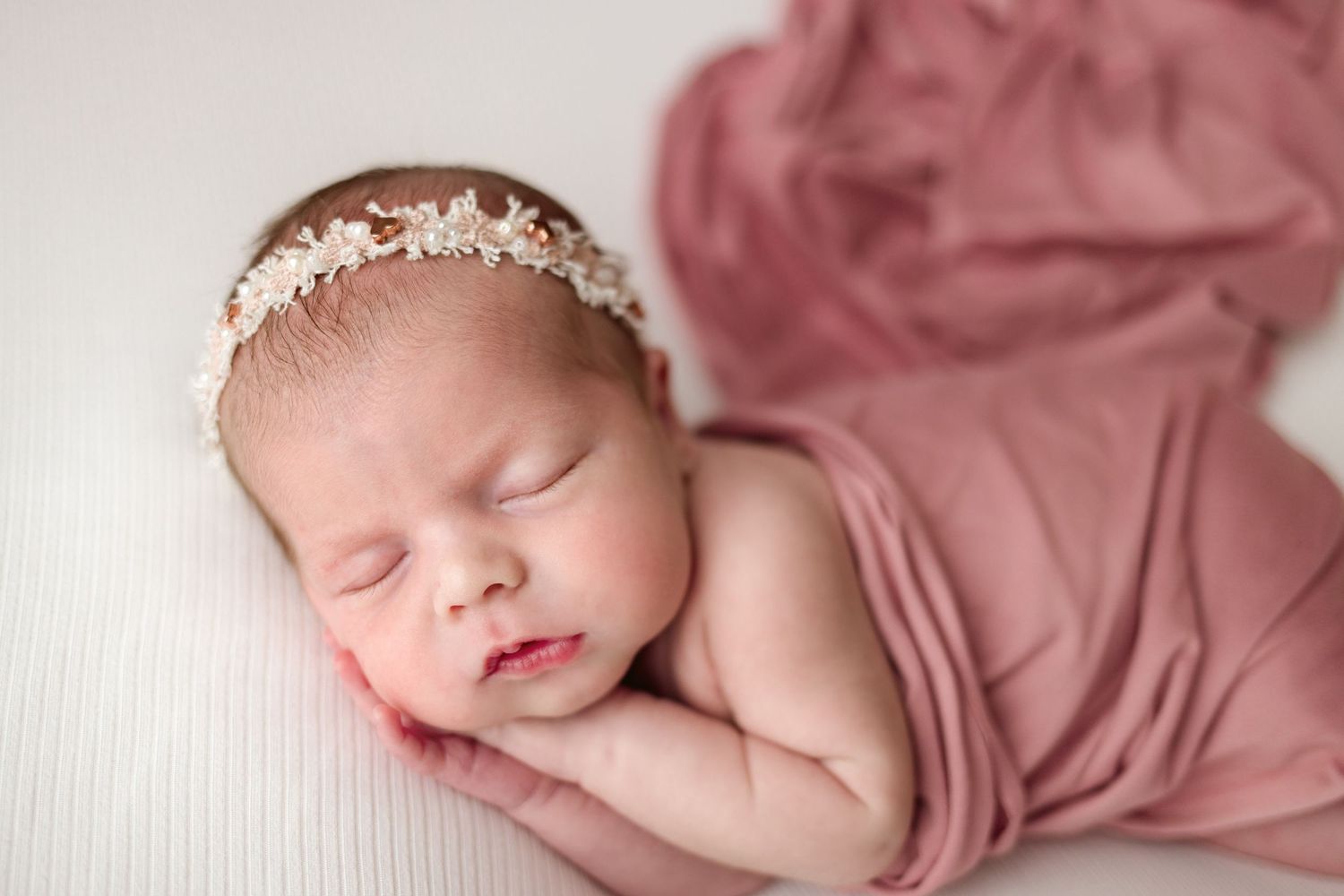 Sleeping newborn baby wrapped in soft pink fabric with delicate lace headband rests peacefully on textured backdrop.