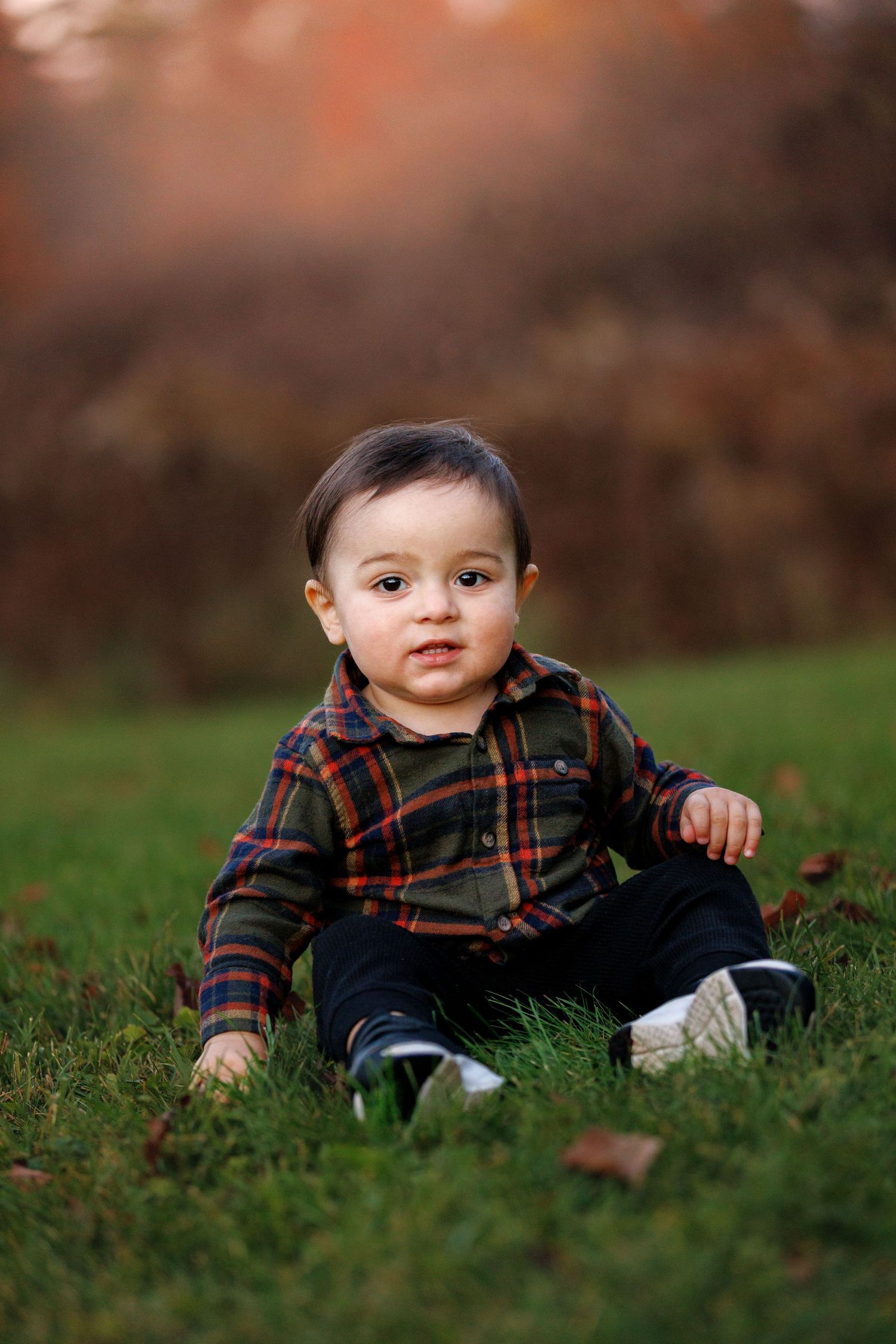 A baby in a plaid shirt lies on grass in an autumn setting during an outdoor photo session.