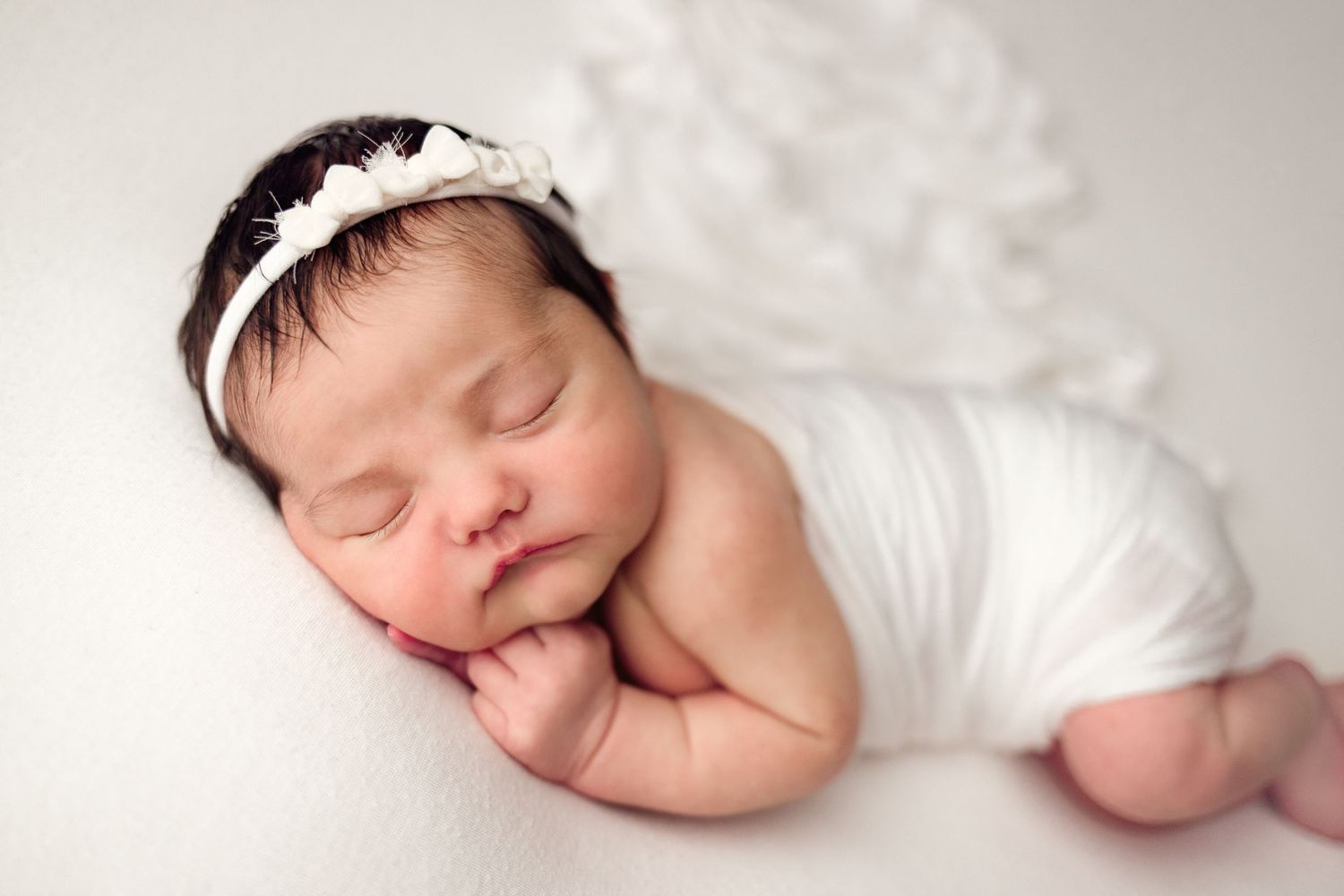 Sleeping newborn baby wearing a white lace headband rests peacefully on soft white bedding.