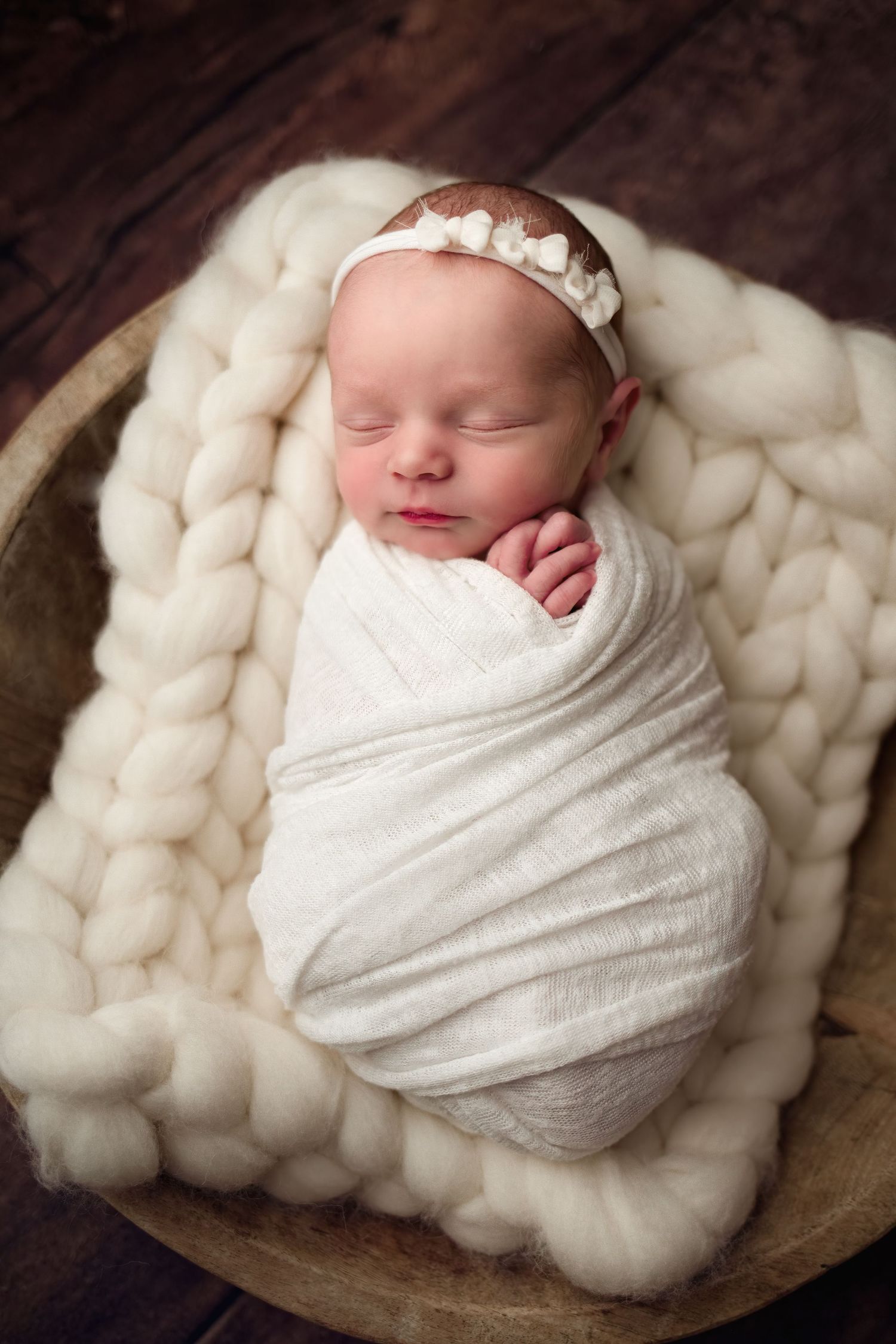 Sleeping newborn baby swaddled in white wrap resting on chunky knit wool blanket in wooden bowl.