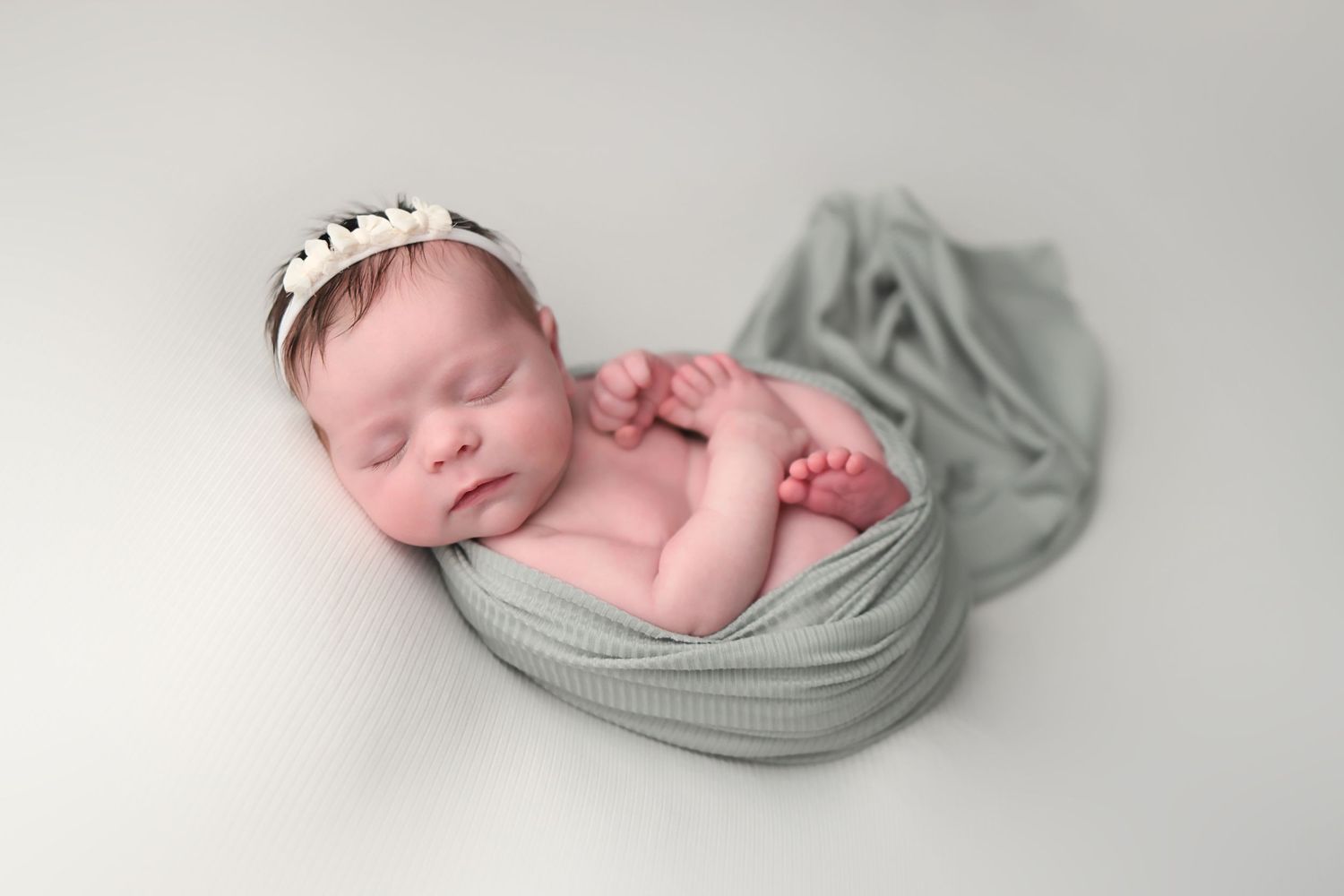 A sleeping newborn baby wrapped in soft sage green fabric lies peacefully in a minimalist photography setup.