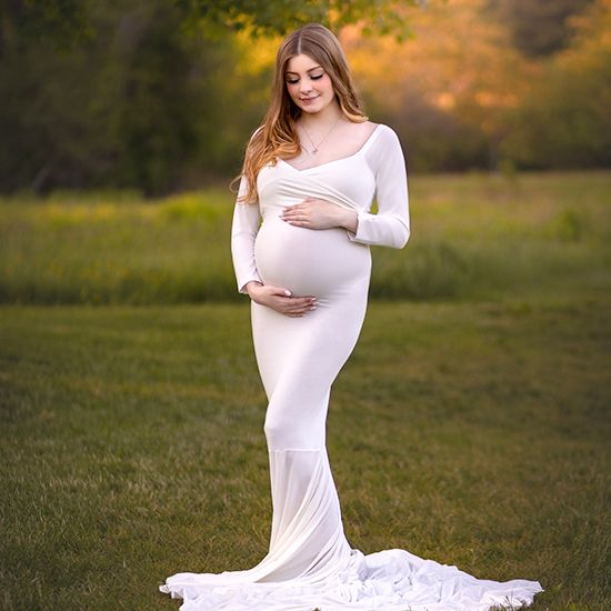 Maternity photoshoot in a white flowing dress against a golden sunset backdrop in a grassy field.