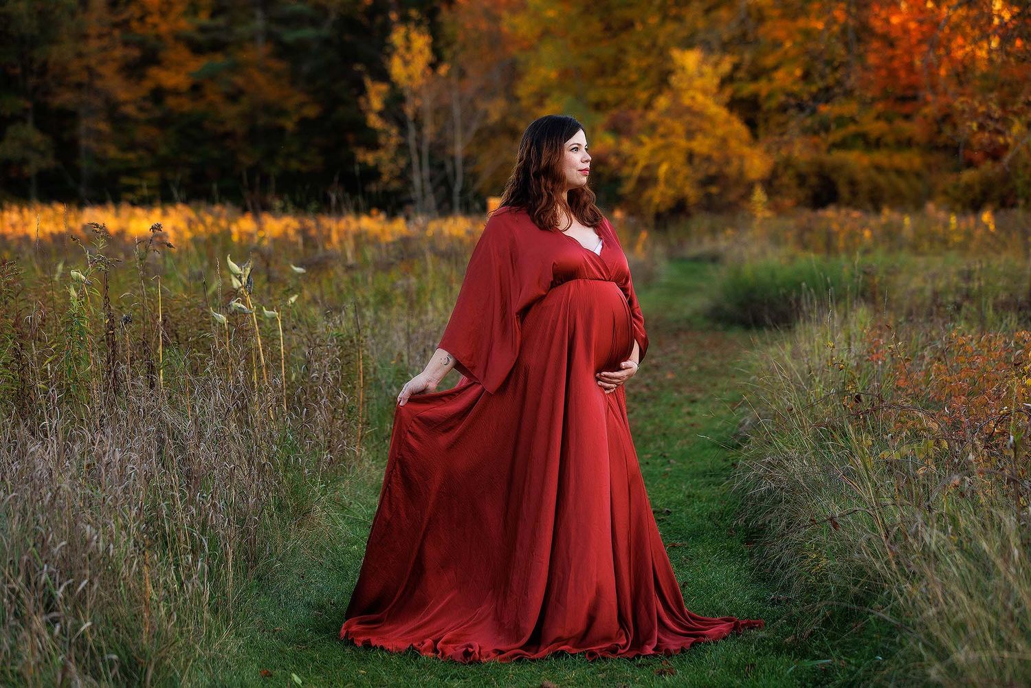 A figure in a flowing burgundy dress poses gracefully in a golden autumn field at sunset.