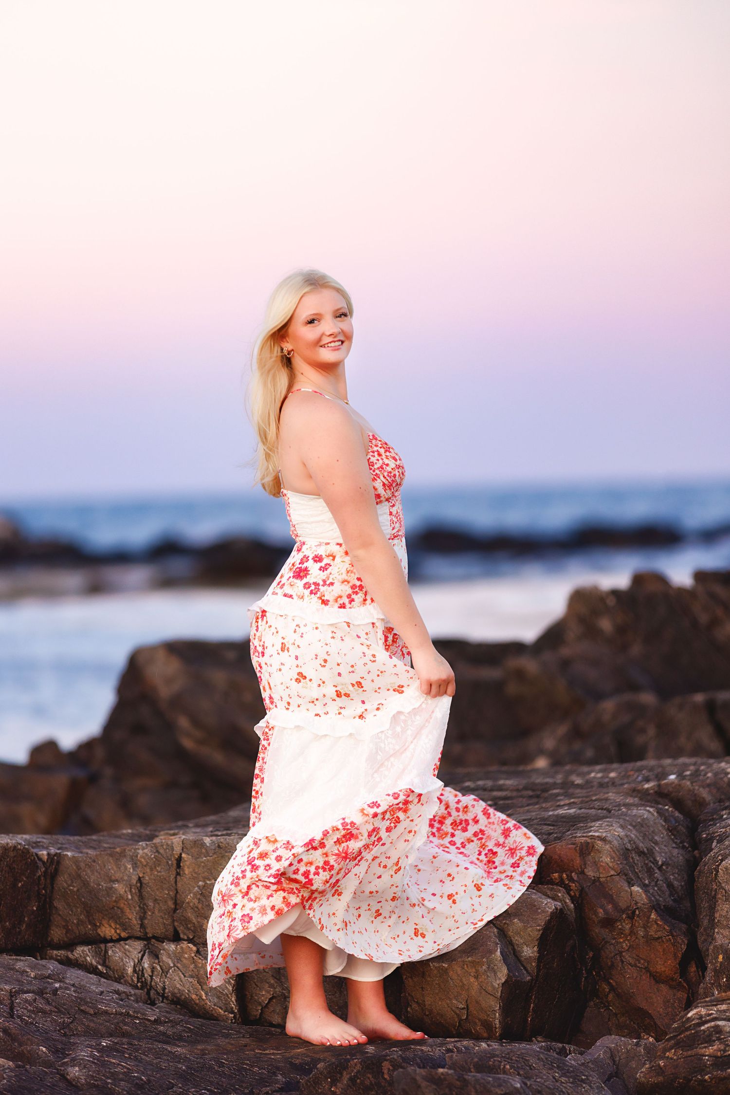 A person in a white and red floral dress twirls on rocky coastal beach at sunset.