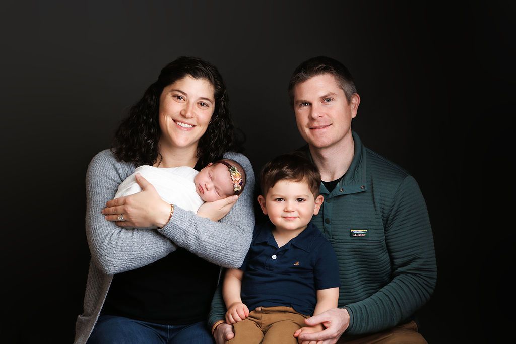 Family portrait with mother holding newborn, toddler son, and father posing together against dark studio background.
