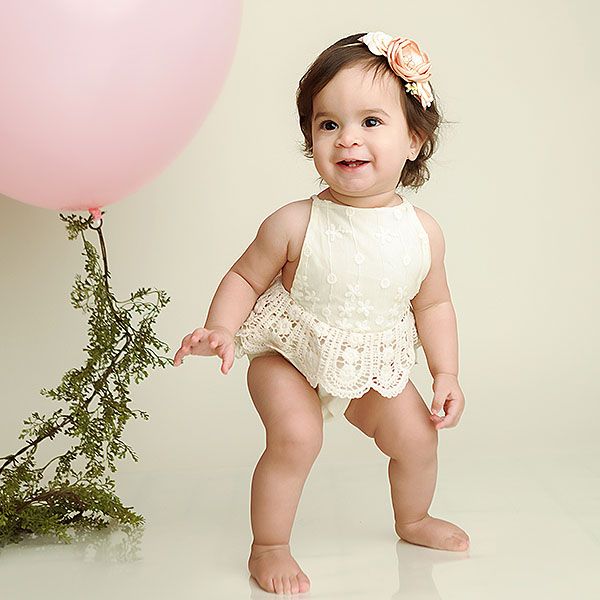 A baby in a white lace dress poses with pink balloons and greenery for a first birthday photo session.