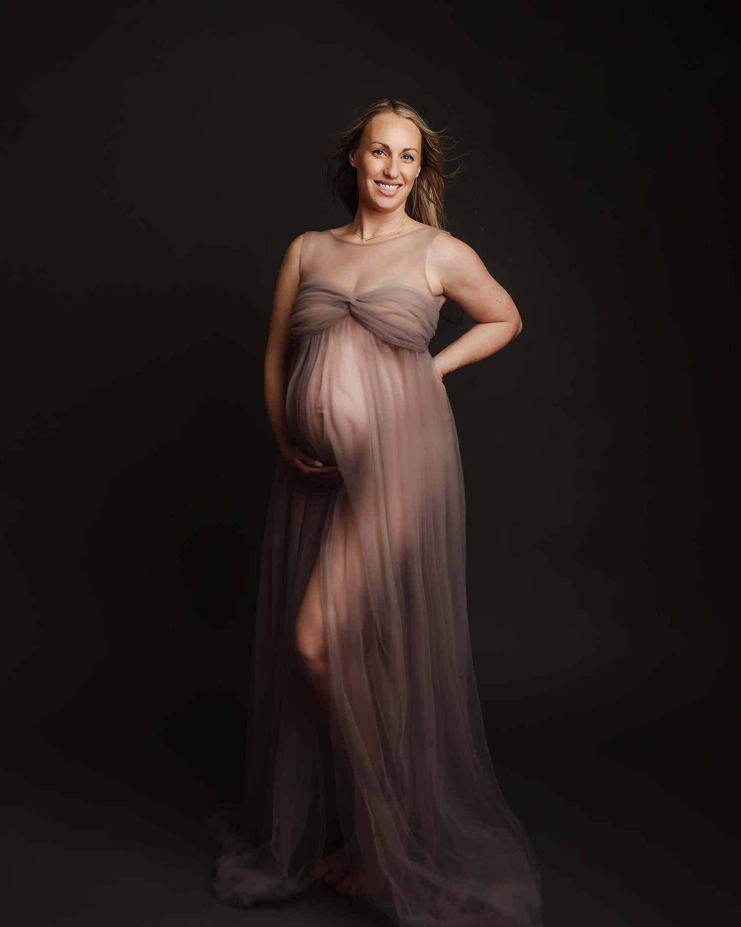A pregnant woman poses in a flowing mauve gown against a dark background in an elegant maternity portrait.