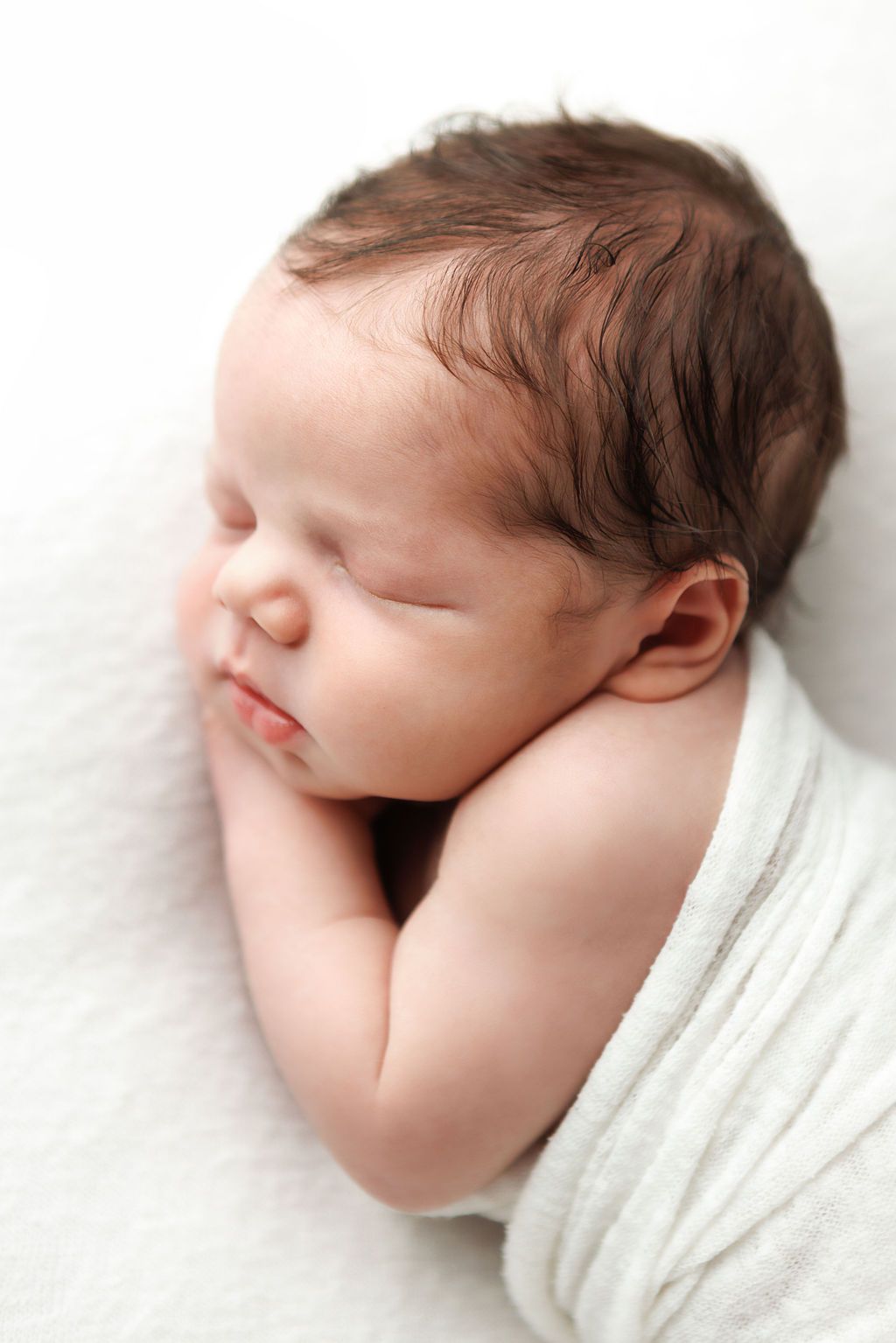 Peaceful newborn baby sleeping wrapped in white blanket with dark hair, serene infant portrait photography.