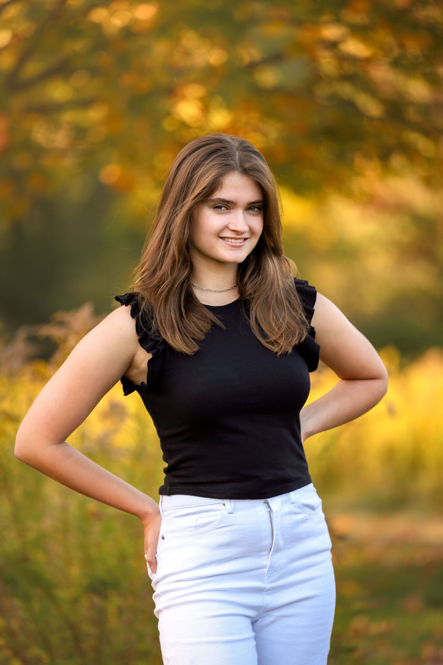 High School Senior in a black top and light pants poses outdoors against a warm autumn background with golden bokeh.