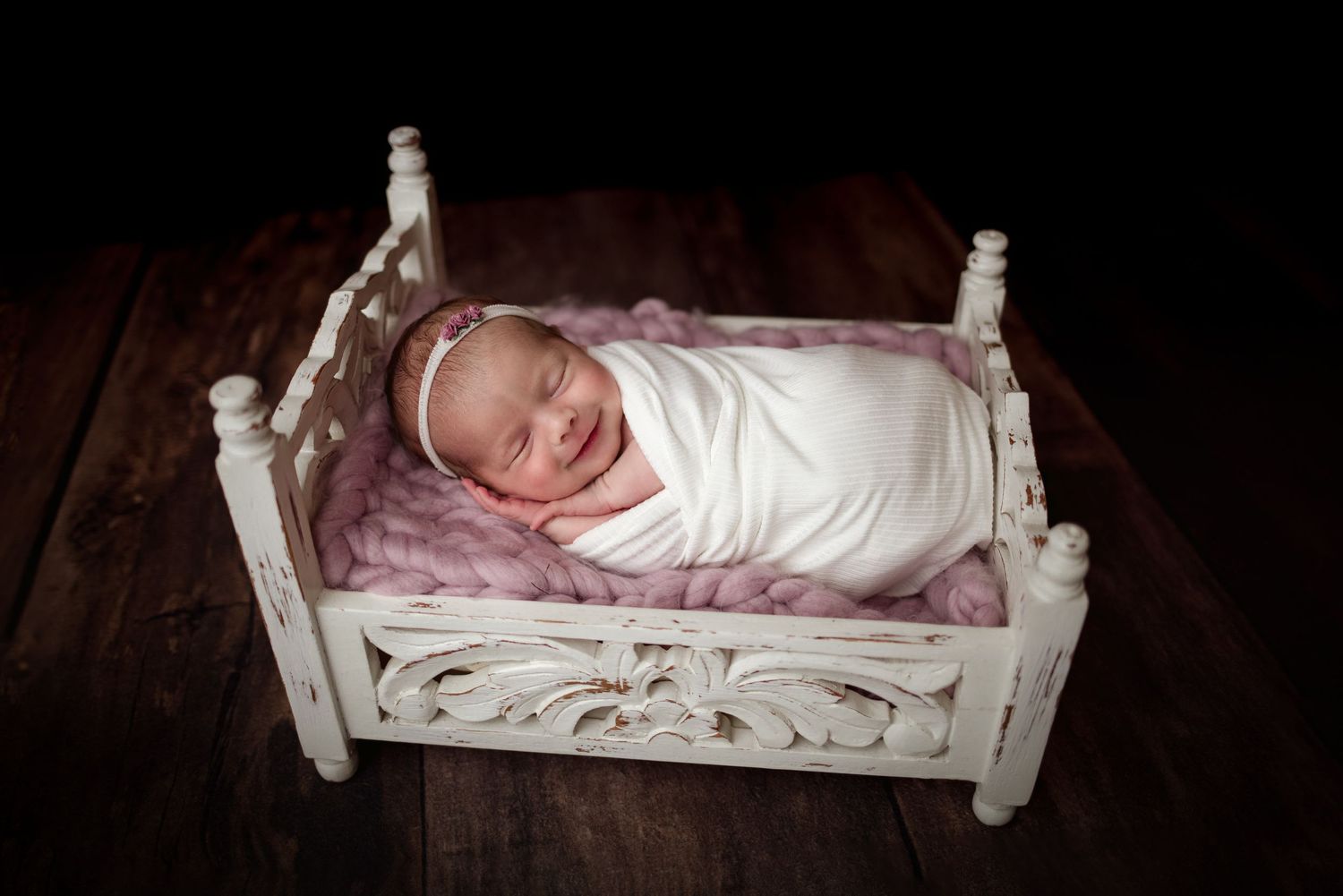 Sleeping newborn baby wrapped in white fabric lying on a small decorative white wooden bed prop.