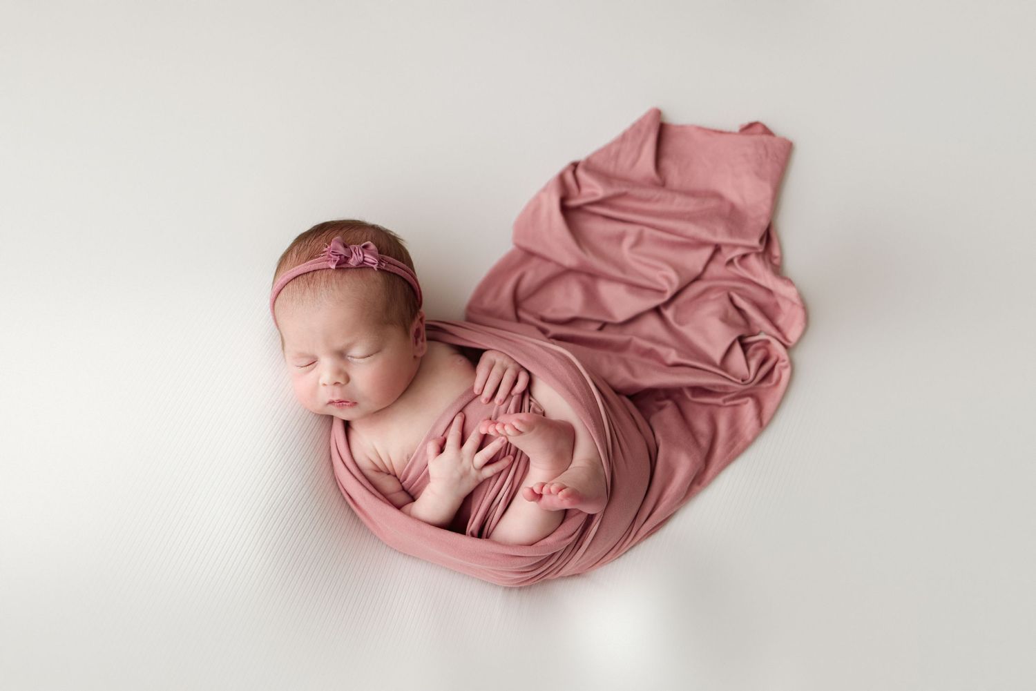 A sleeping newborn baby wrapped in soft dusty rose fabric on a white background.