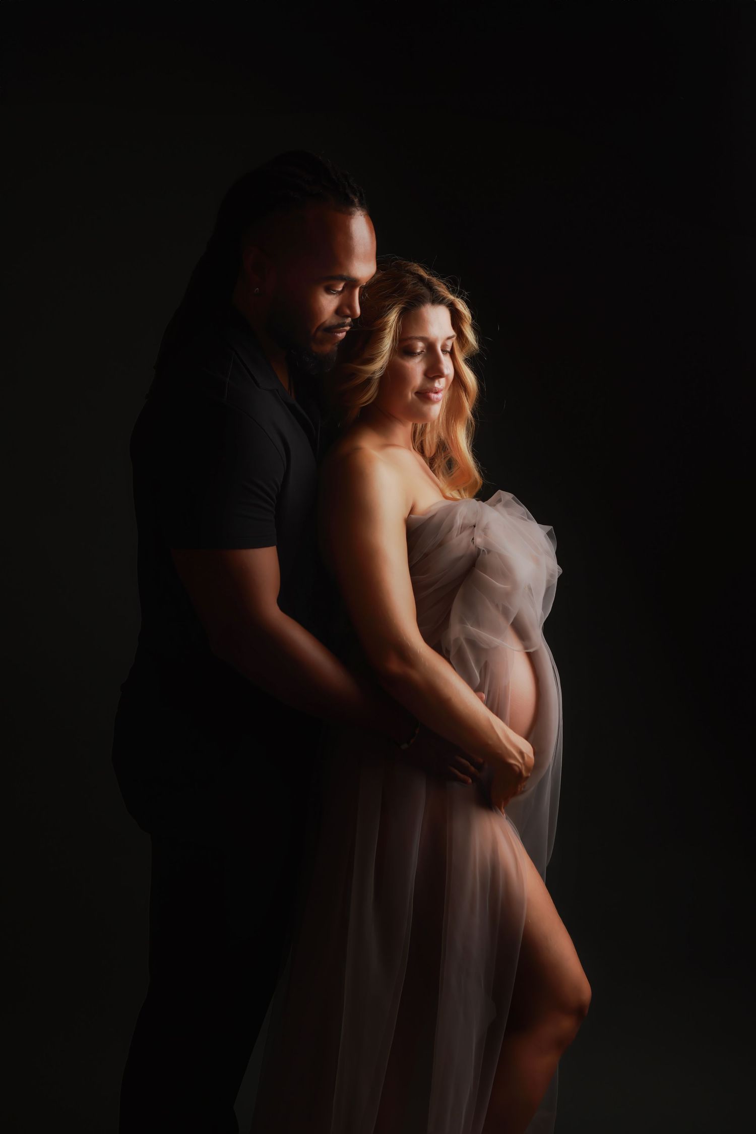 Intimate maternity portrait featuring couple in dramatic lighting with flowing sheer dress against dark background.