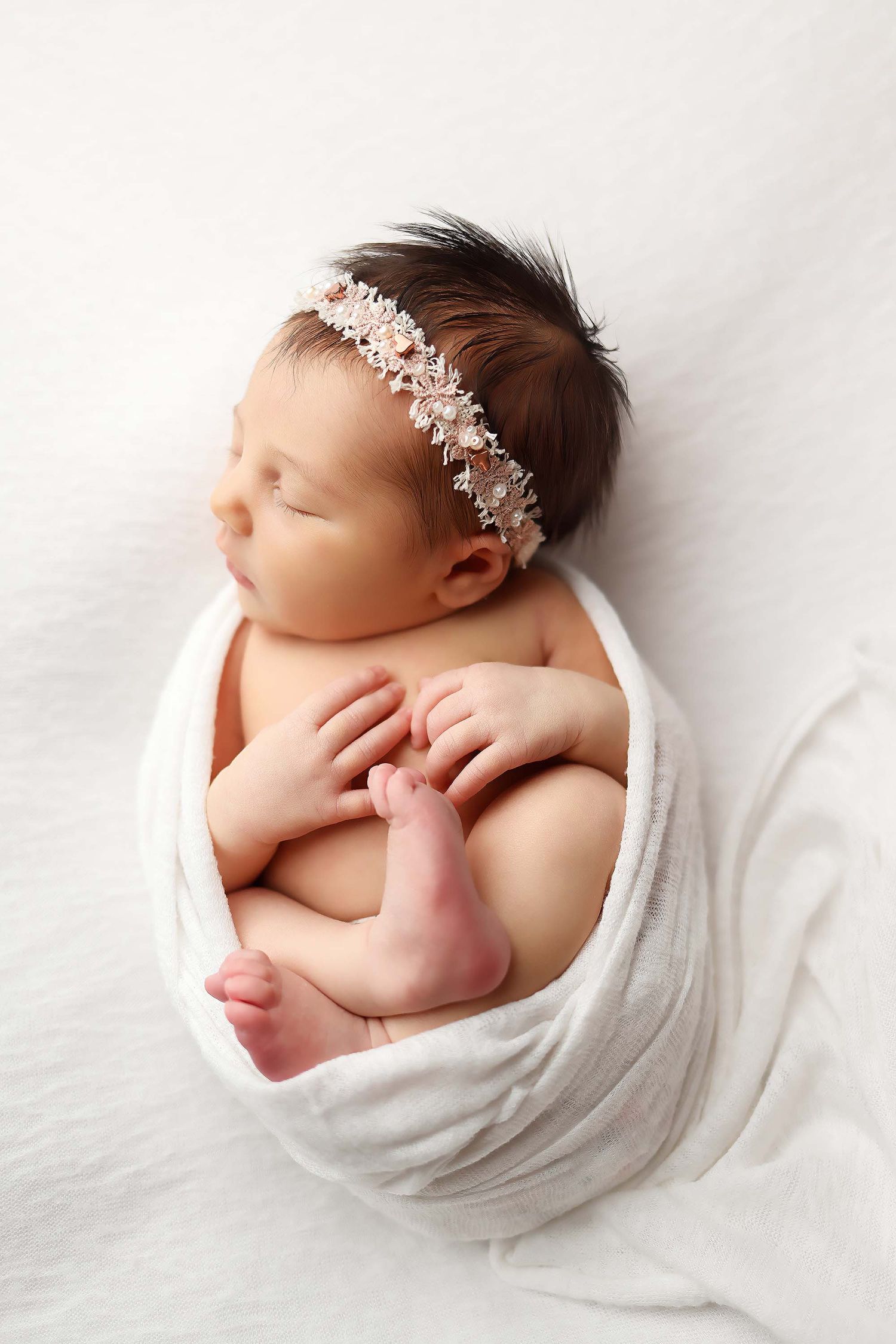Sleeping newborn wrapped in white fabric with delicate floral headpiece on white background.