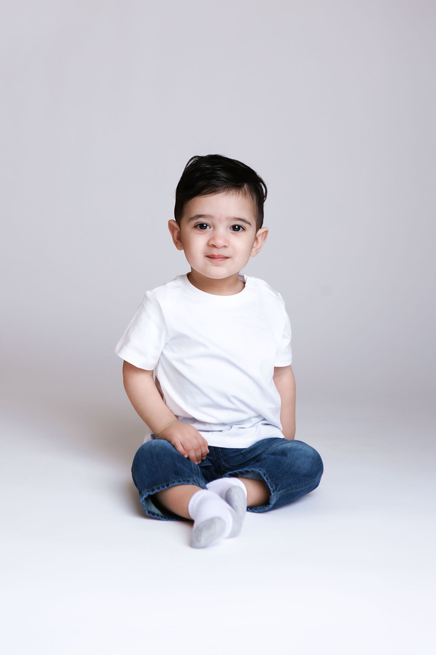 Cute toddler boy sitting cross-legged in white t-shirt and jeans, smiling at camera on white background.