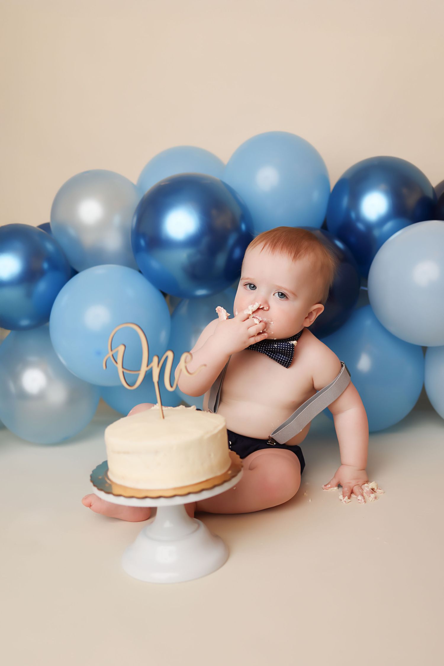 Baby boy celebrates first birthday smashing a white cake, surrounded by blue balloons and wearing a bow tie.