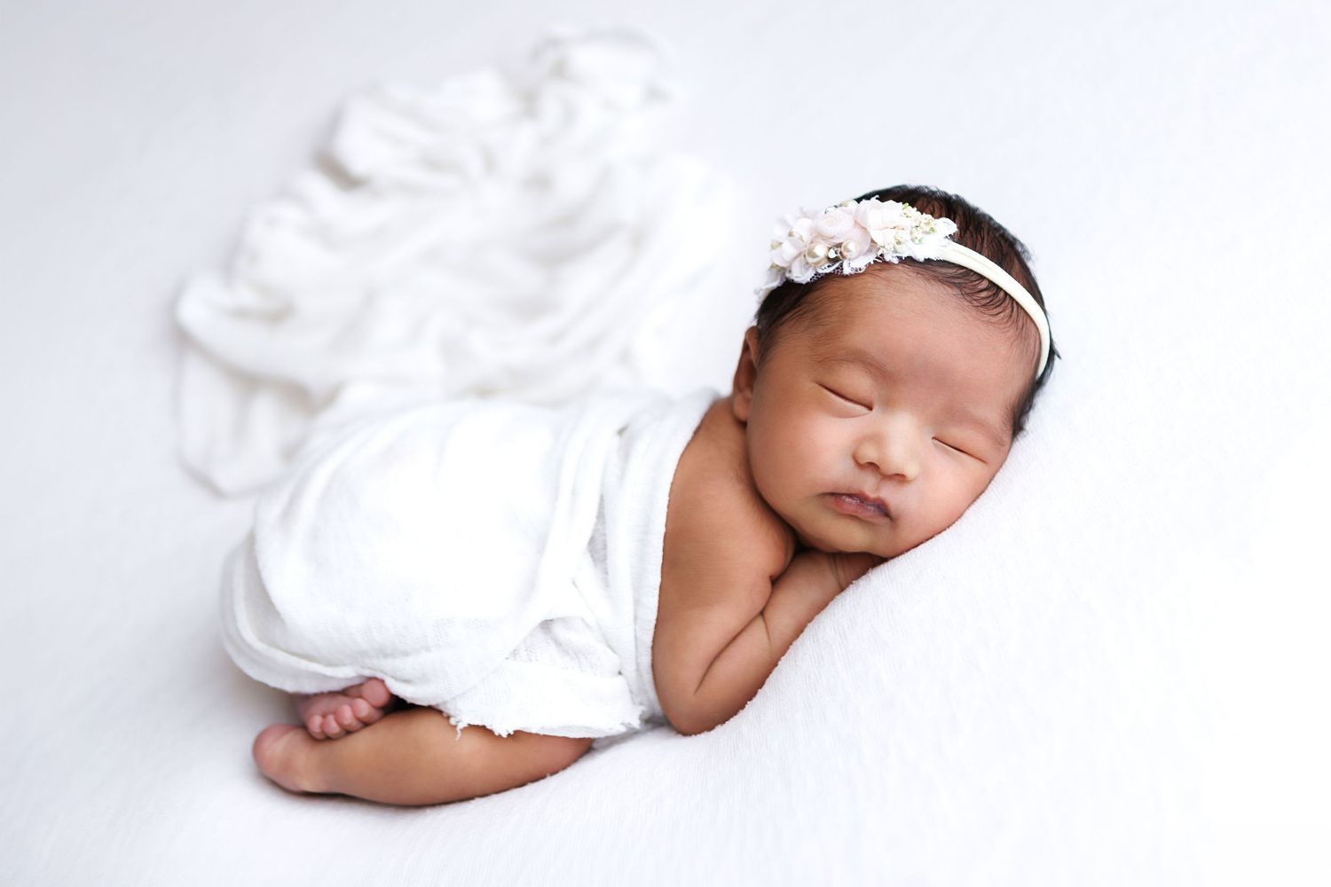 Sleeping newborn baby wrapped in white cloth with floral headband on white background.