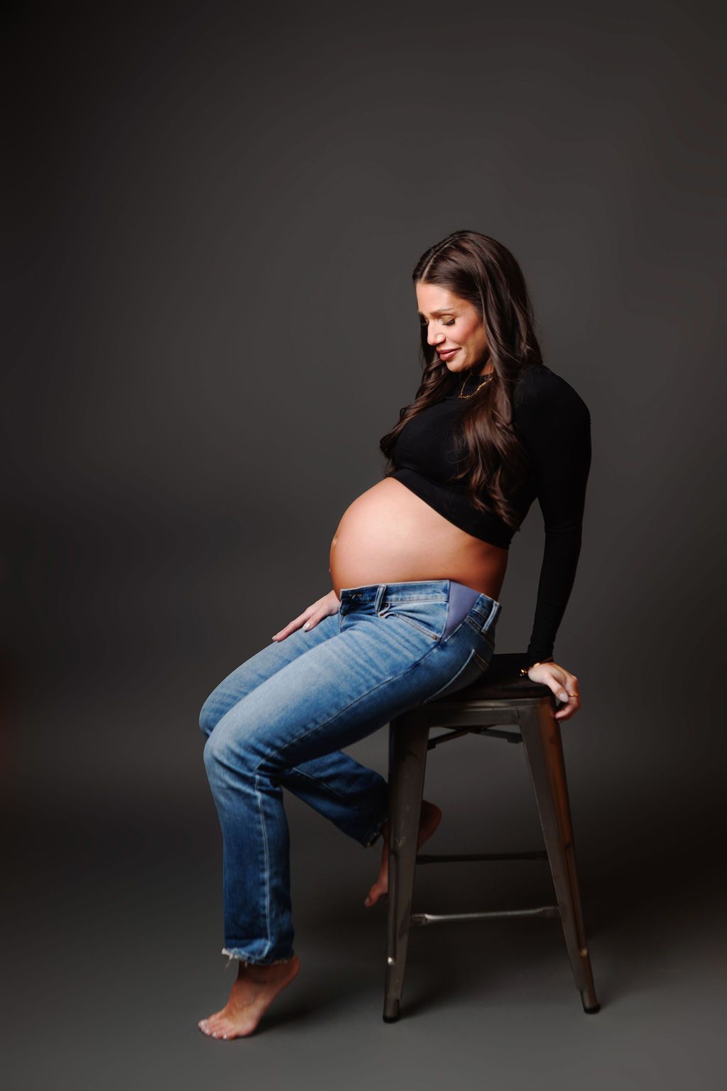 Pregnant woman sitting on a stool in black top and jeans, cradling her bare baby bump in a studio.